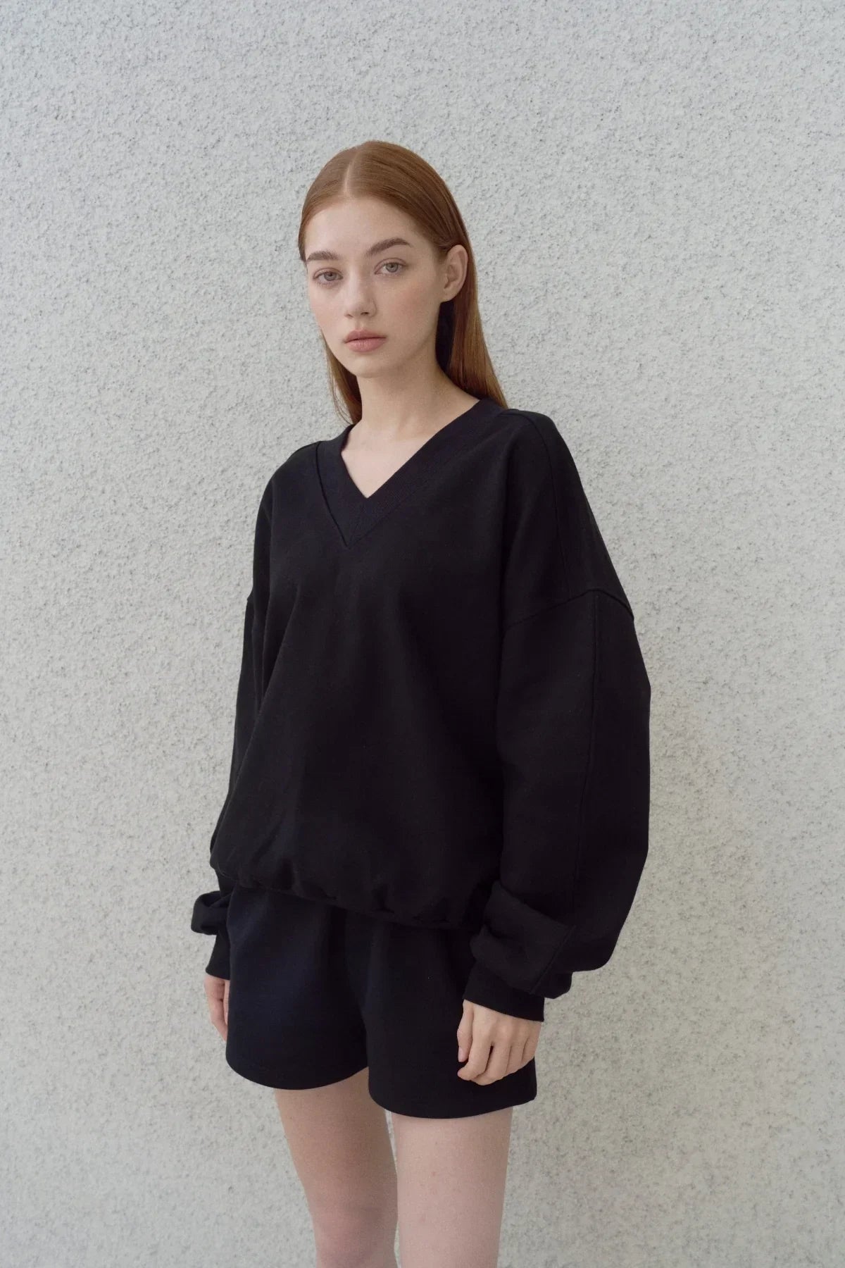 Model wearing black heavy cotton V-neck sweatshirt and black shorts against gray textured wall