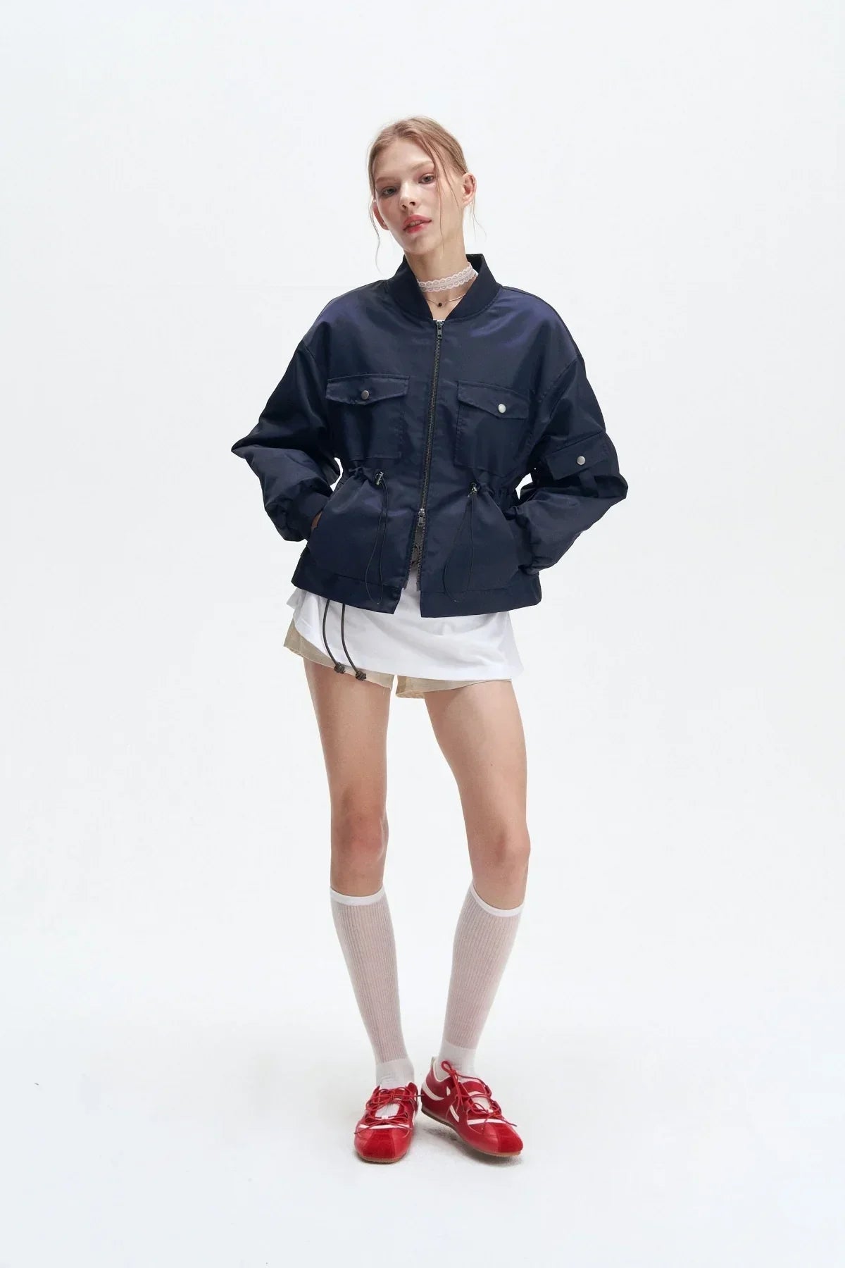 Model wearing a navy heavy nylon bomber jacket with pockets, white skirt, knee-high socks, and red shoes