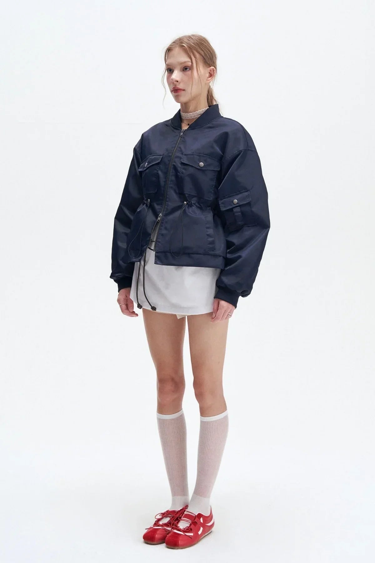 Model wearing navy heavy nylon bomber jacket with elastic cuffs, white skirt, knee-high socks, and red sneakers