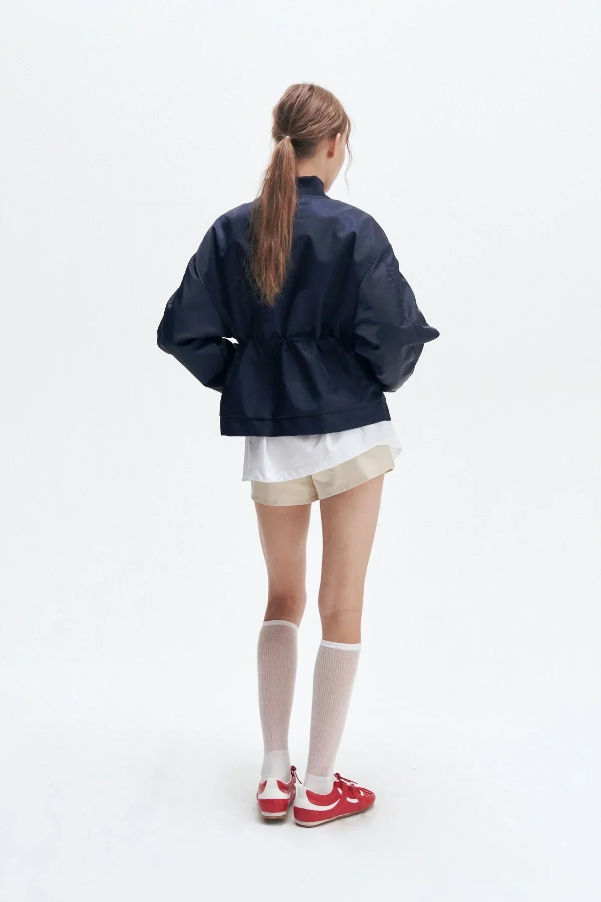 Woman wearing navy heavy nylon bomber jacket with beige shorts, white knee-high socks, and red sneakers