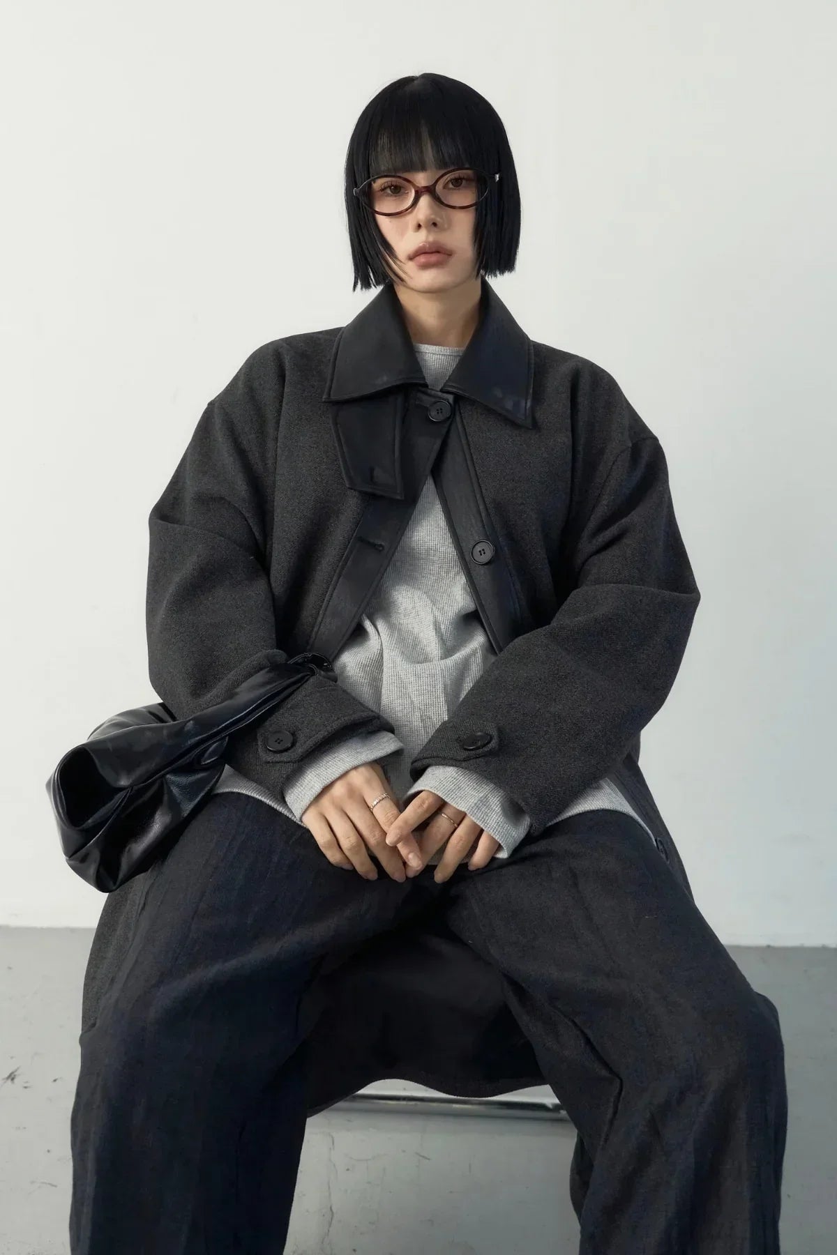 Woman with short black hair and glasses wearing a dark oversized high-neck long coat and gray sweater