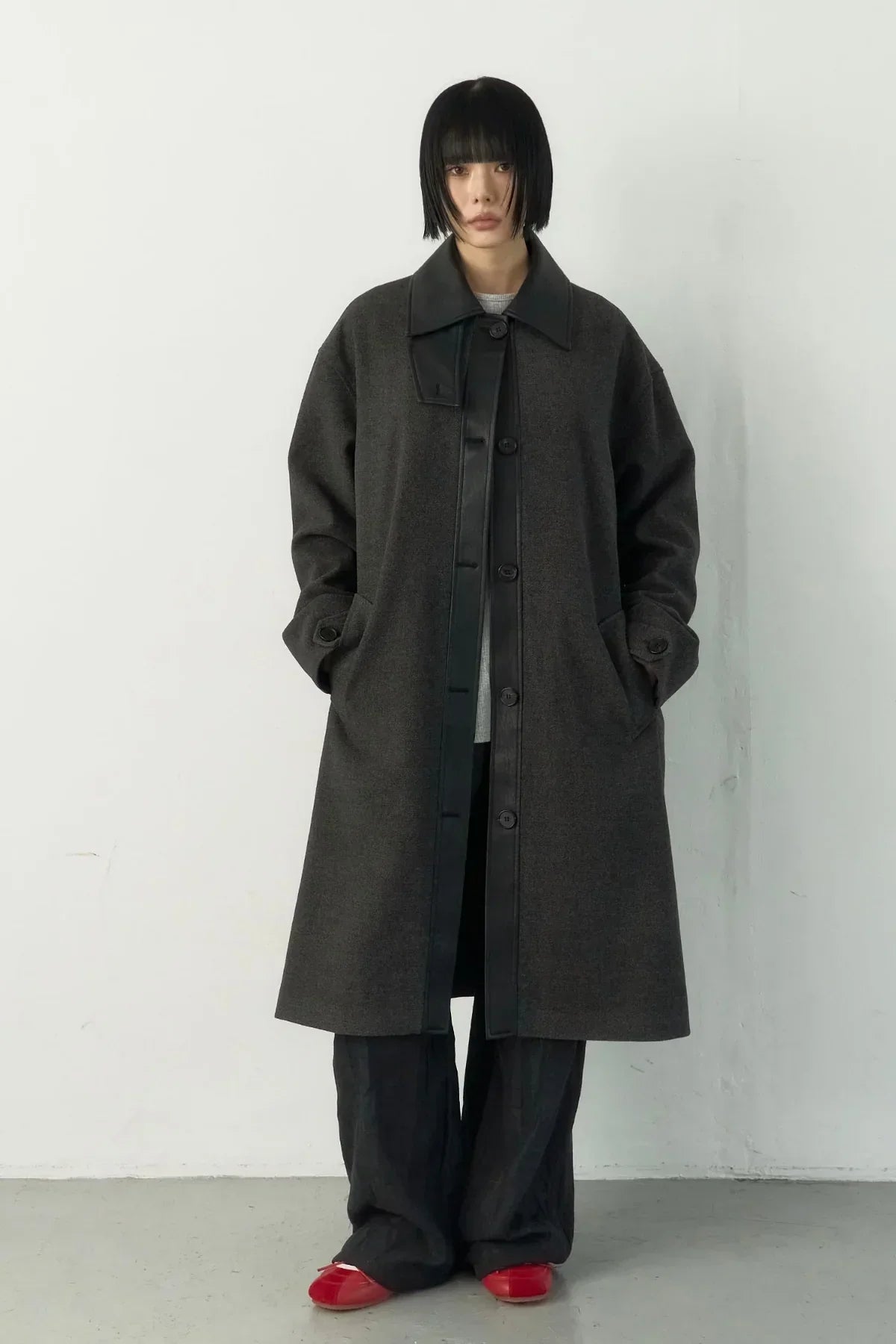 Model wearing dark grey long single-breasted coat with black trim, wide pants, and red shoes in studio