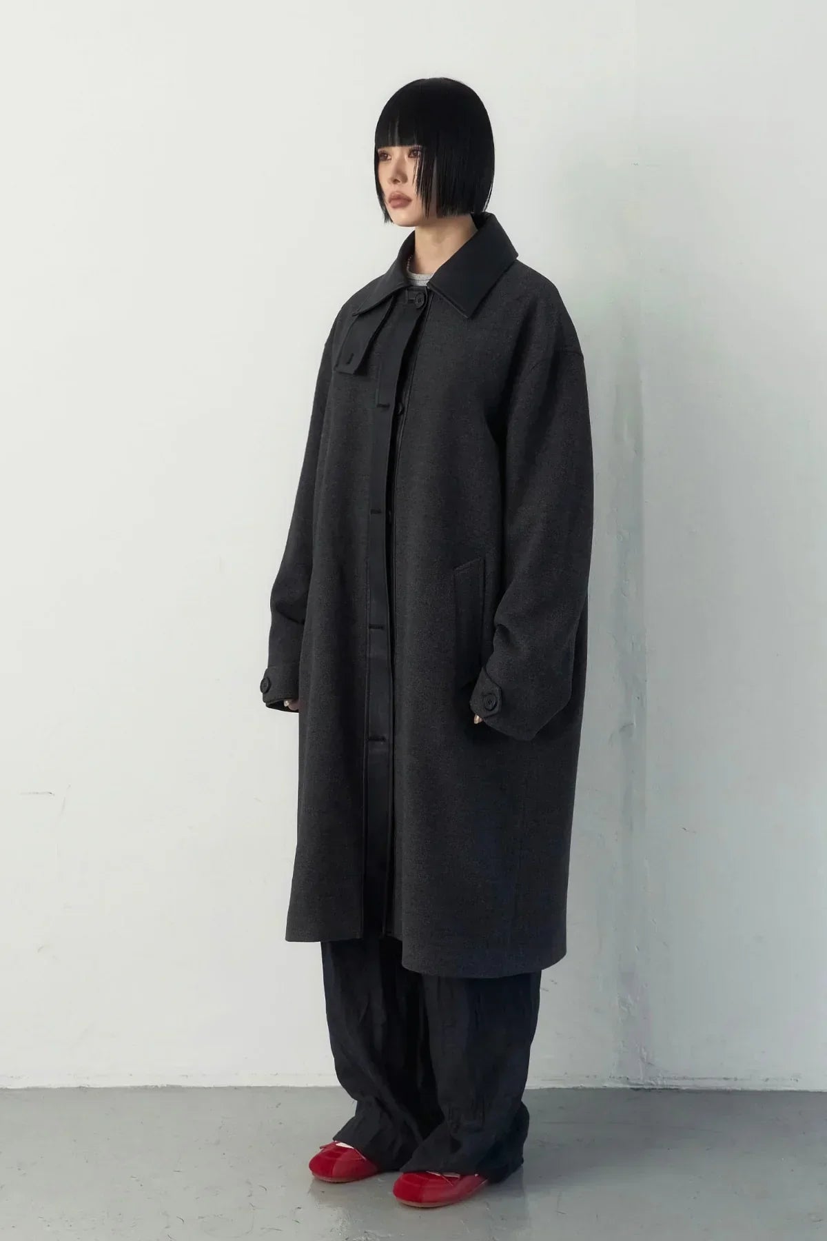 Model wearing a high-neck charcoal gray long coat, loose black pants, and red shoes against a plain white wall
