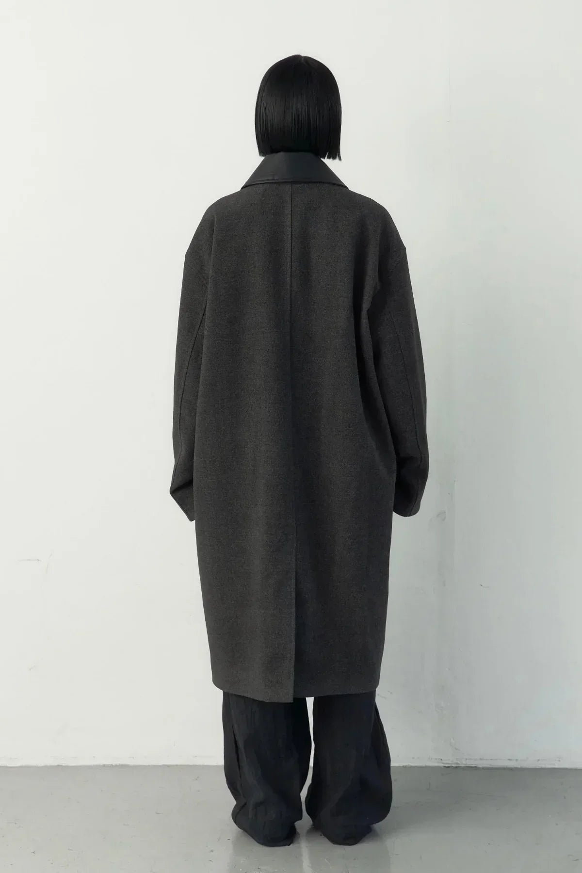 Back view of model wearing a dark gray high-neck long single coat with black wide-leg pants