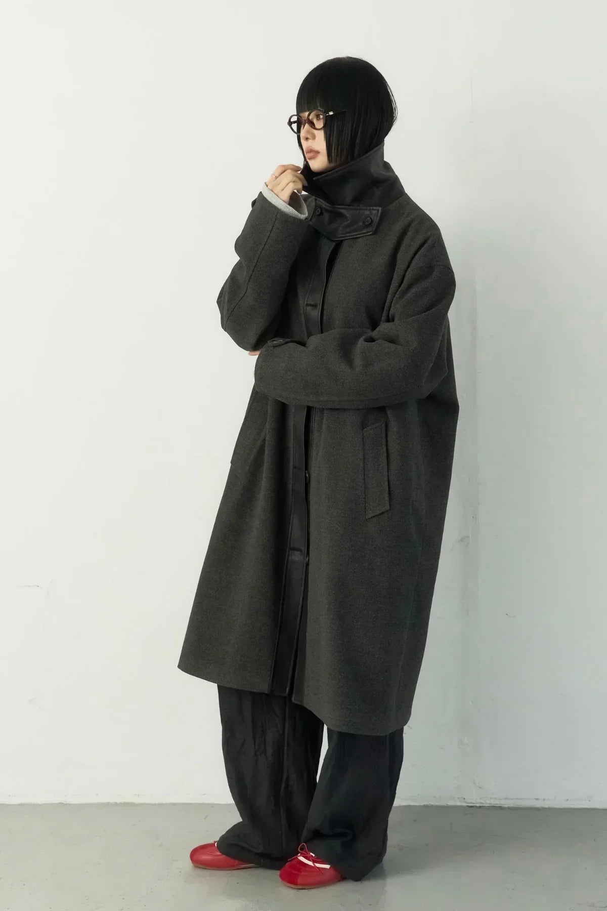 Model wearing a dark gray high-neck long coat with black wide pants and red shoes in studio