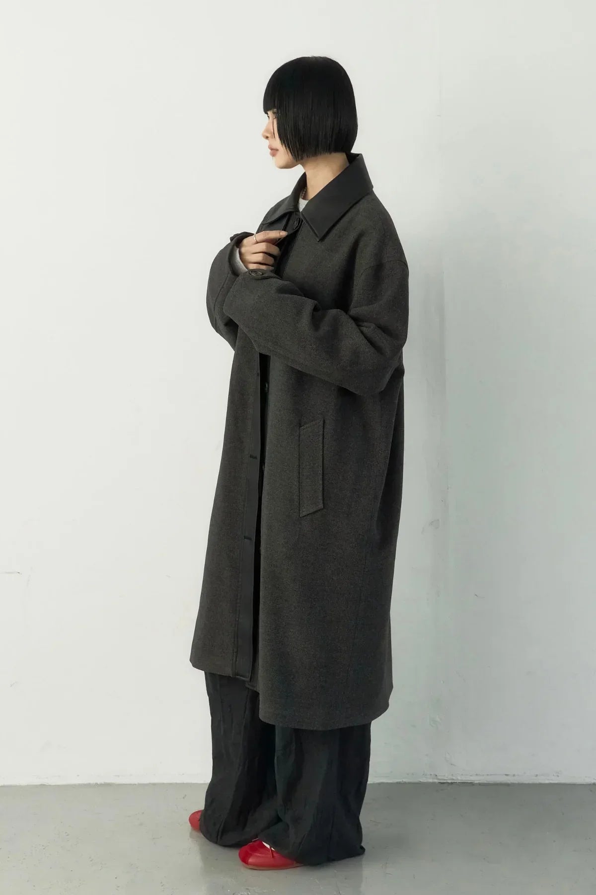 Woman in dark oversized long coat with large collar, black wide pants, and red shoes against white wall