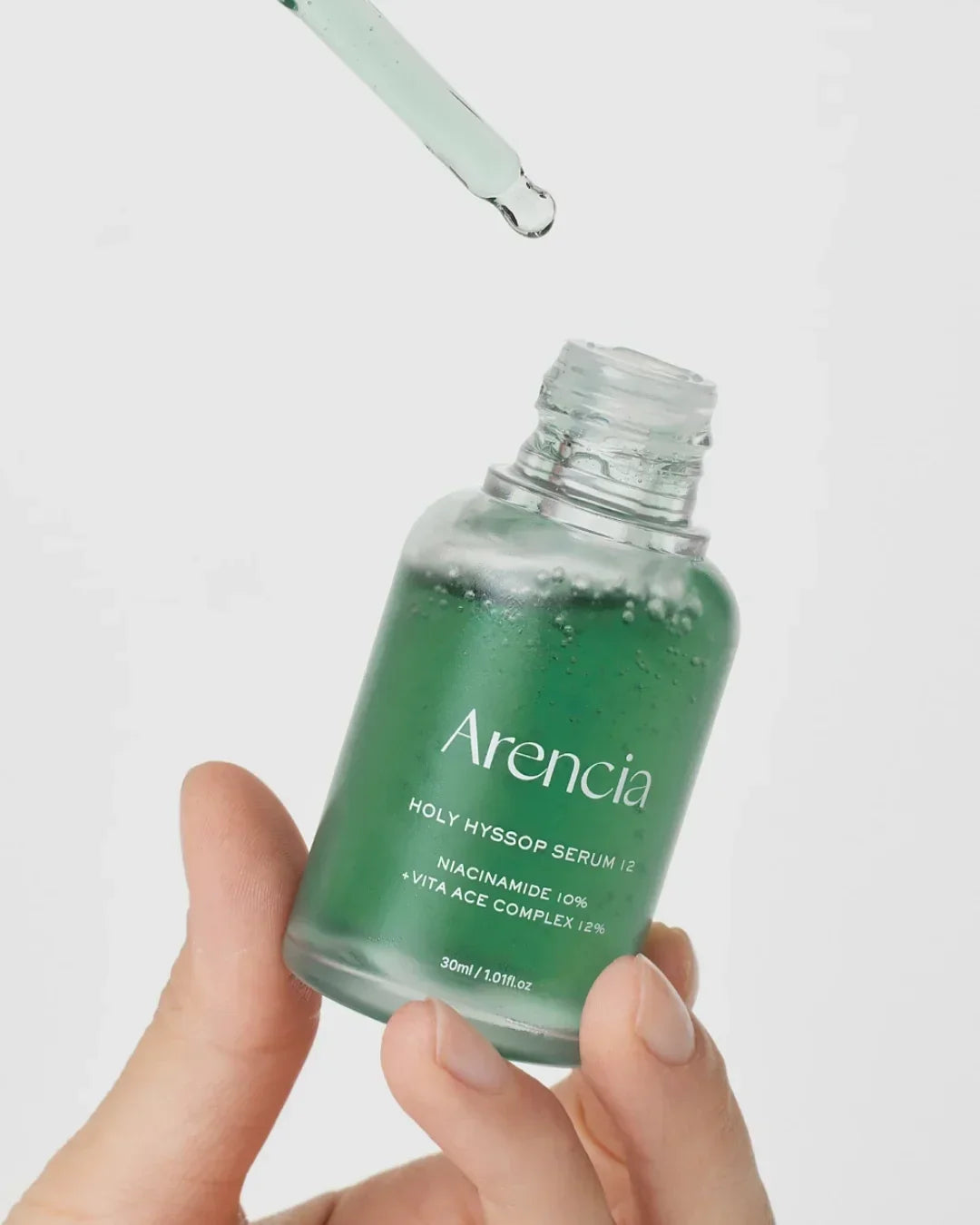Hand holding open green serum bottle labeled Arencia Holy Hyssop Serum with dropper releasing liquid