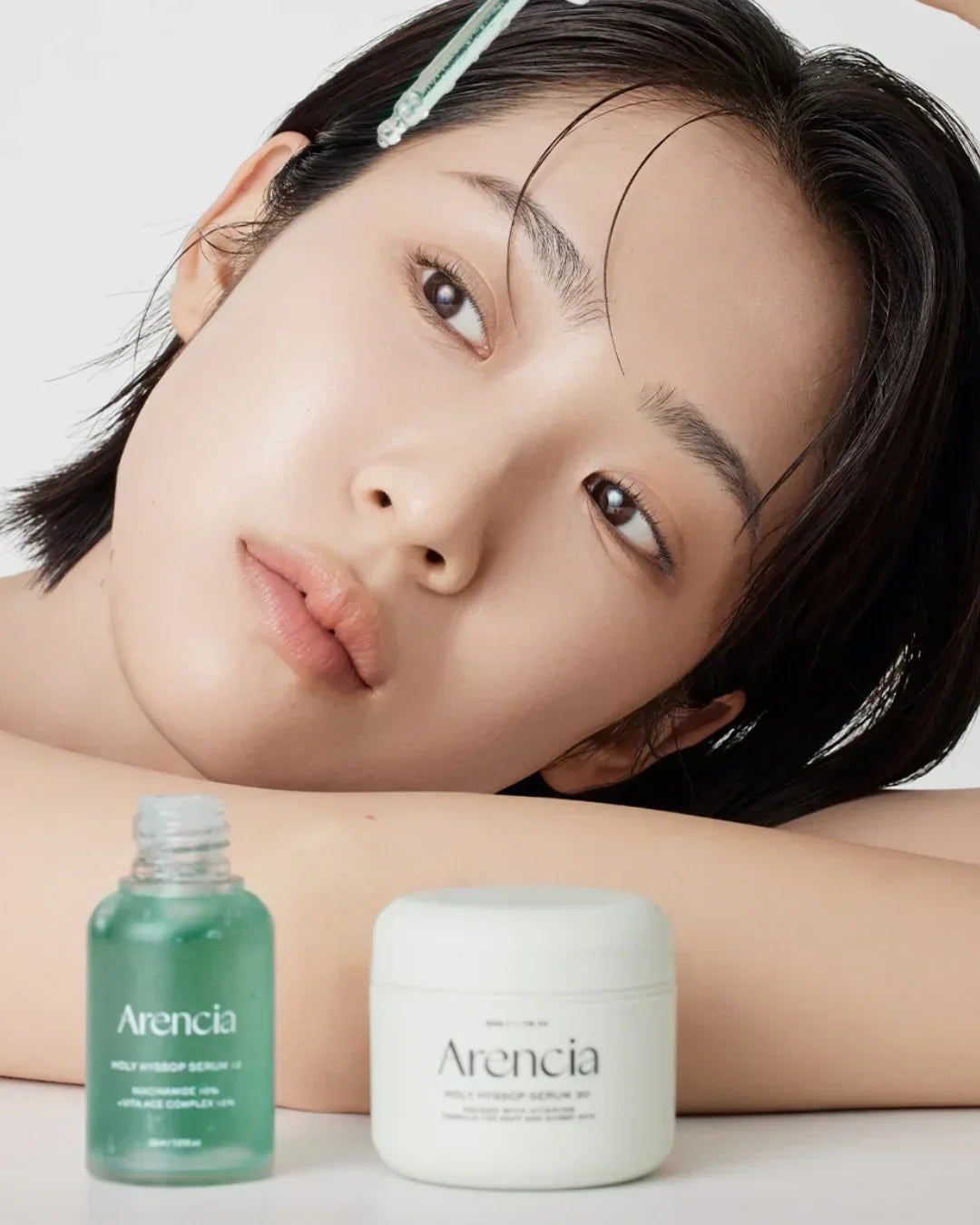 Close-up of woman with clear skin using Arencia Holy Hyssop Serum dropper, serum bottle and cream jar