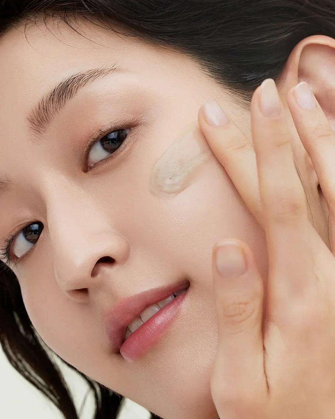Close-up of woman applying creamy white skincare serum on cheek for radiant skin