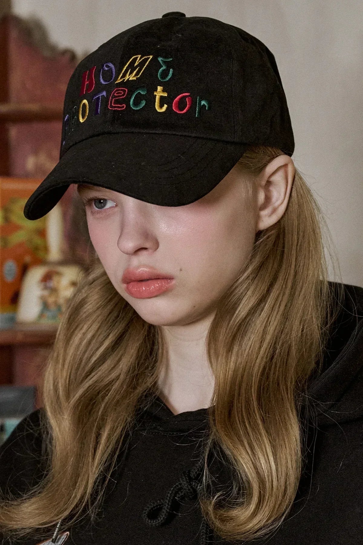 Close-up of a woman with long blonde hair wearing a black 'Home Protector' embroidered ballcap and black hoodie