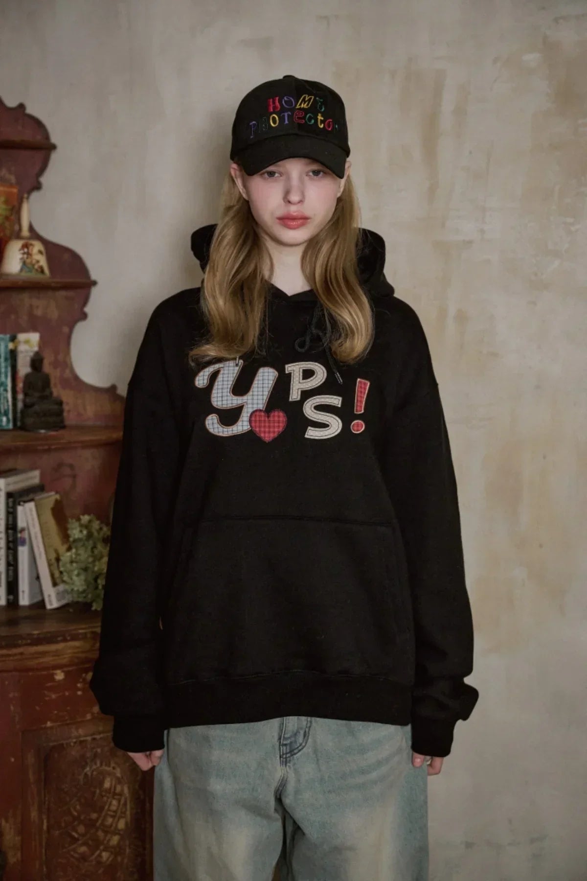 Young woman wearing black hoodie with Yups text and black Home Protector embroidered ballcap in indoor vintage setting