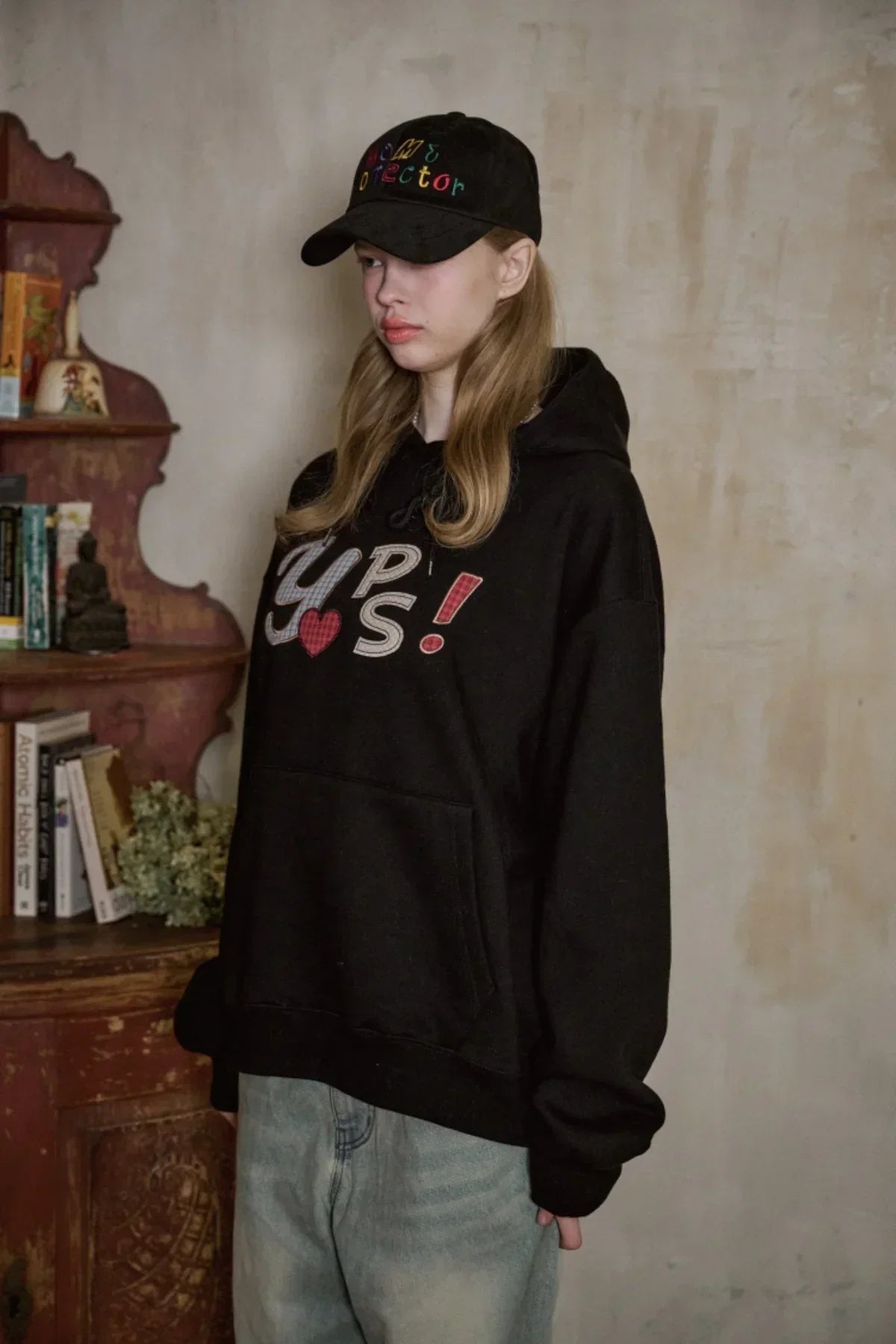 Young woman wearing black Home Protector baseball cap and oversized black hoodie with text, standing indoors