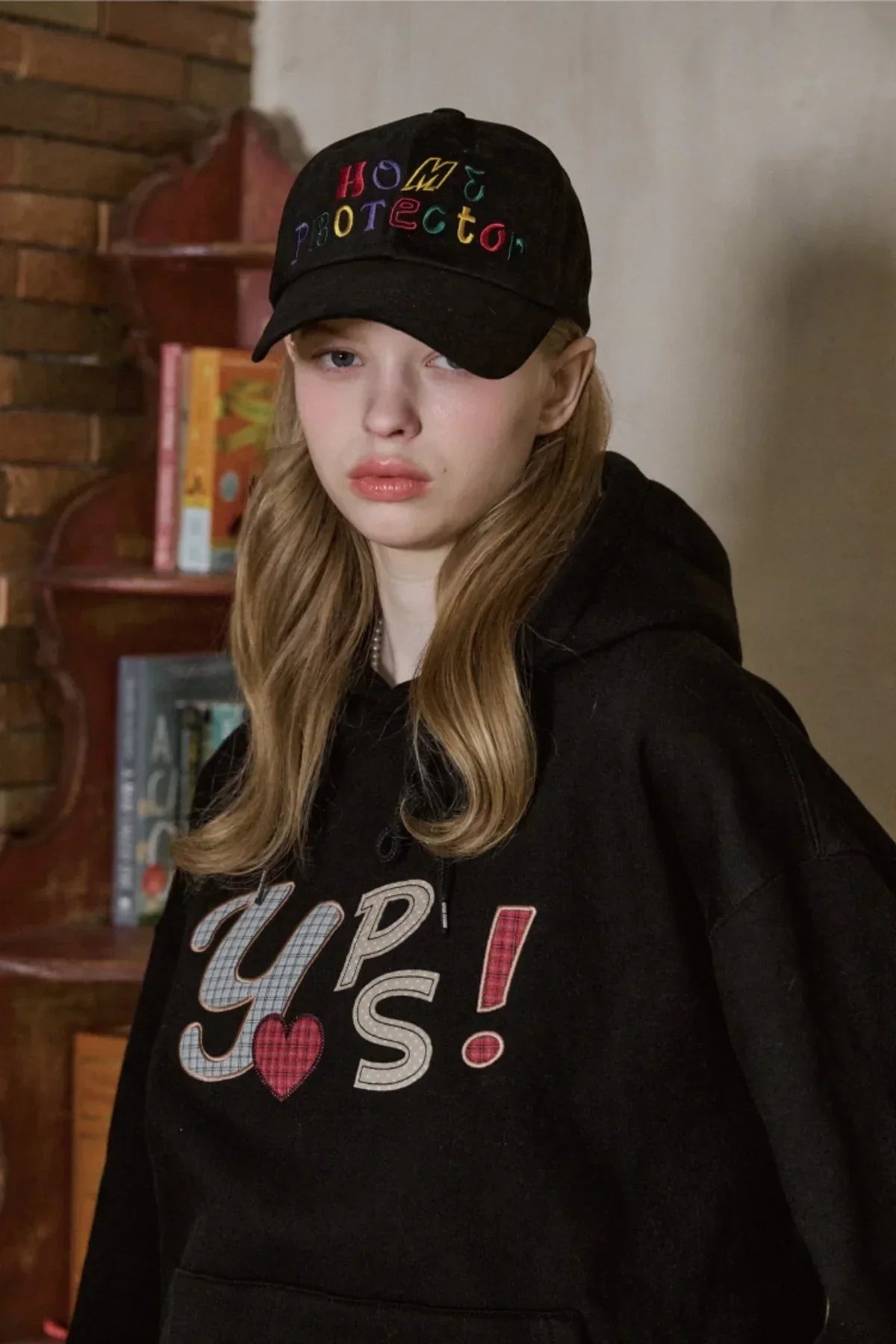 Young woman wearing black Home Protector cap and black hoodie with colorful text in an indoor setting