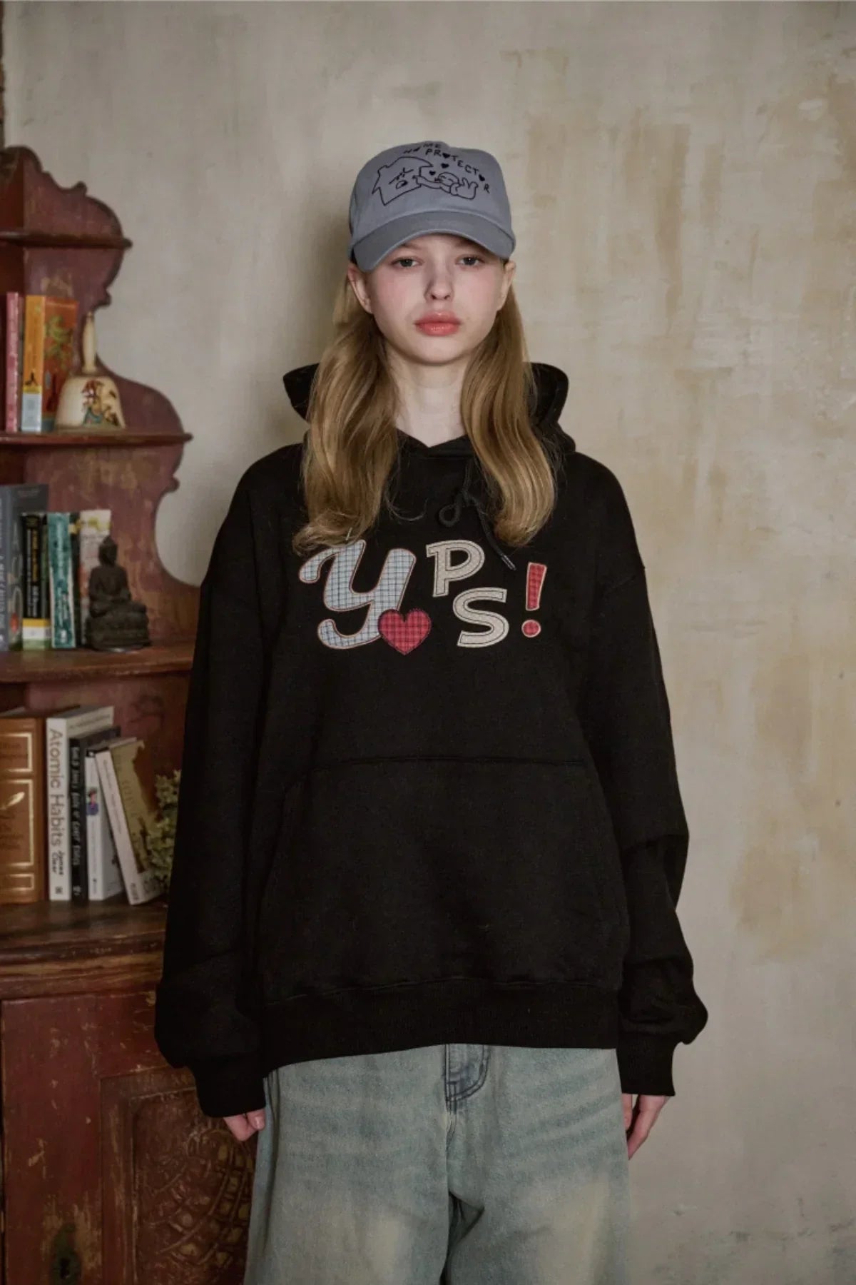 Young woman wearing a black hoodie with 'Y UPS!' text and a sky-blue baseball cap indoors