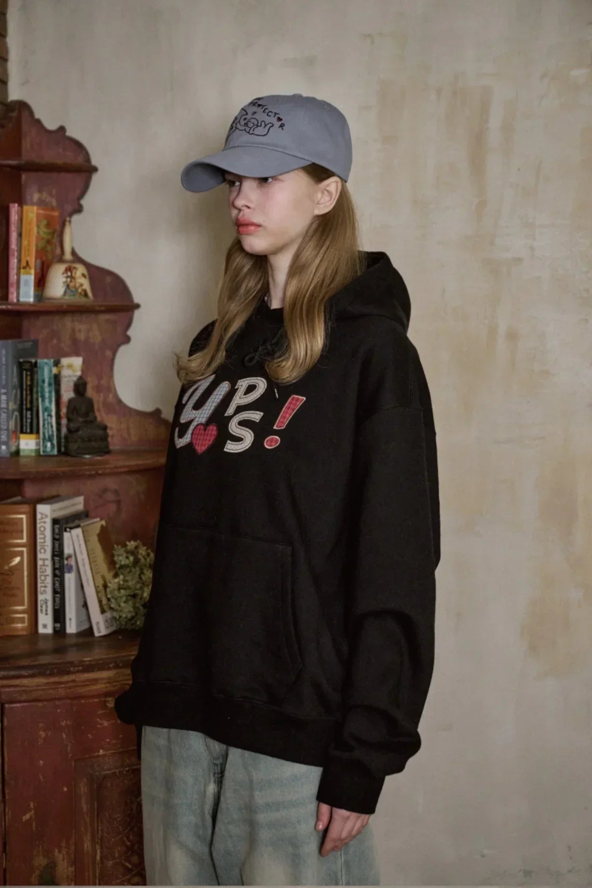 Young woman wearing a black hoodie with graphic text and a blue embroidered ballcap indoors