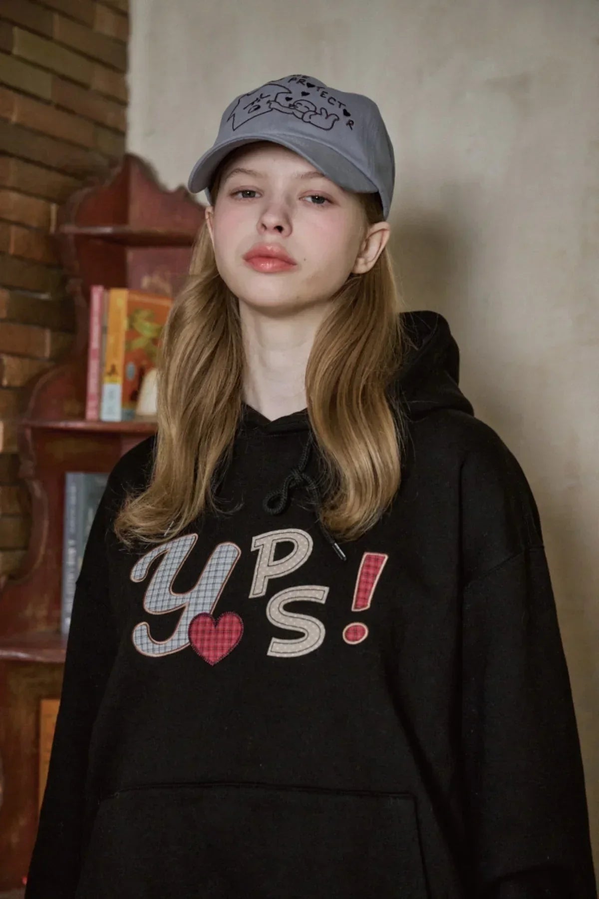 Young woman wearing a black hoodie with colorful 'YPS!' print and a gray ballcap in a cozy indoor setting