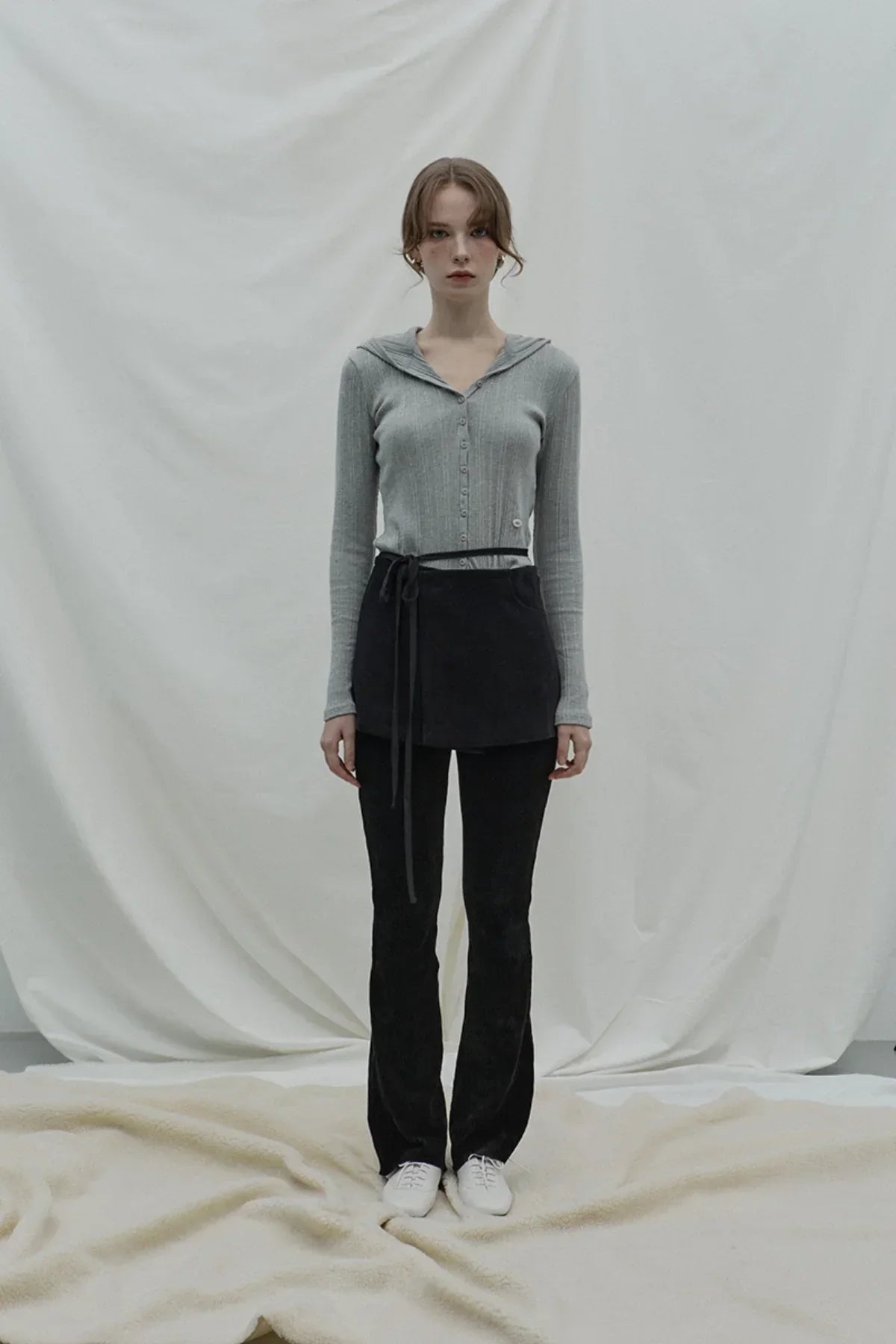 Woman wearing a gray ribbed hooded cardigan with black belted skirt and black pants in minimal studio