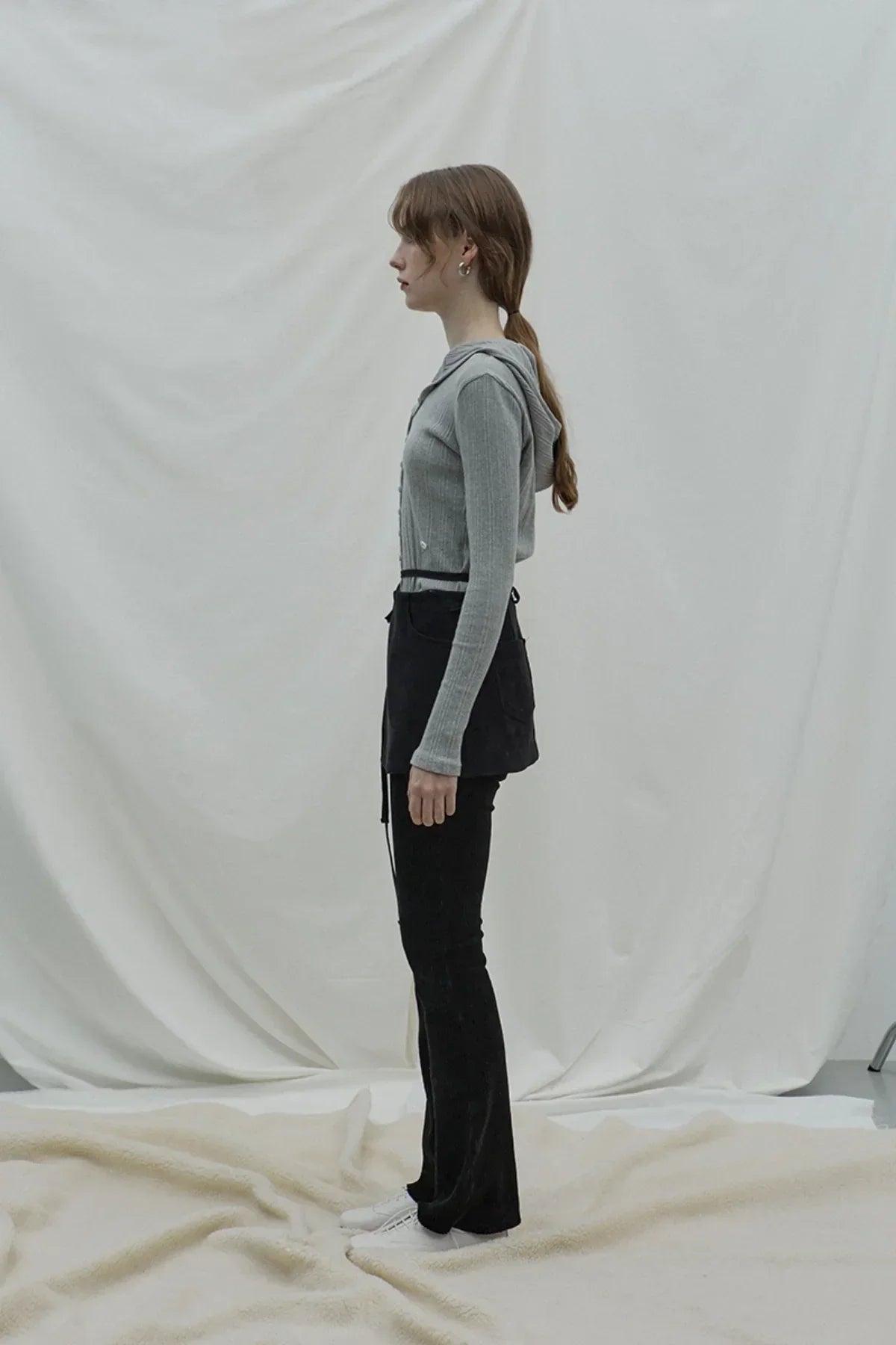 Side view of a woman wearing a gray ribbed hooded cardigan, black skirt, black pants, and white shoes against a white fabric backdrop