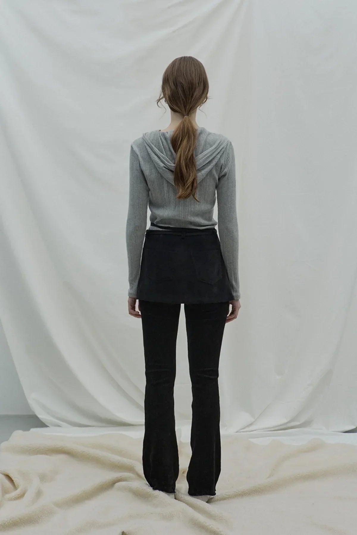 Rear view of woman wearing gray ribbed hooded cardigan and black pants against white backdrop