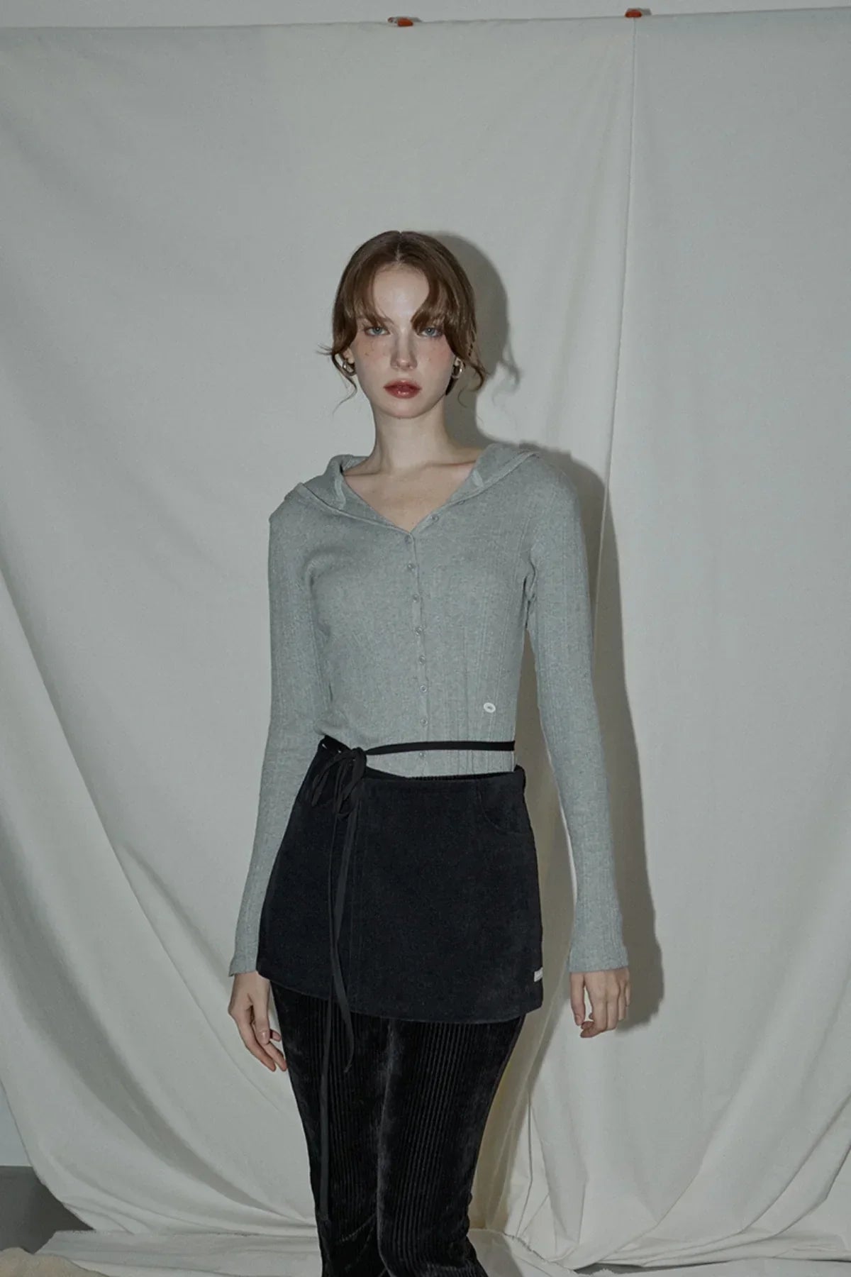 Woman wearing gray ribbed hooded cardigan with button-down front and black skirt over black pants in studio