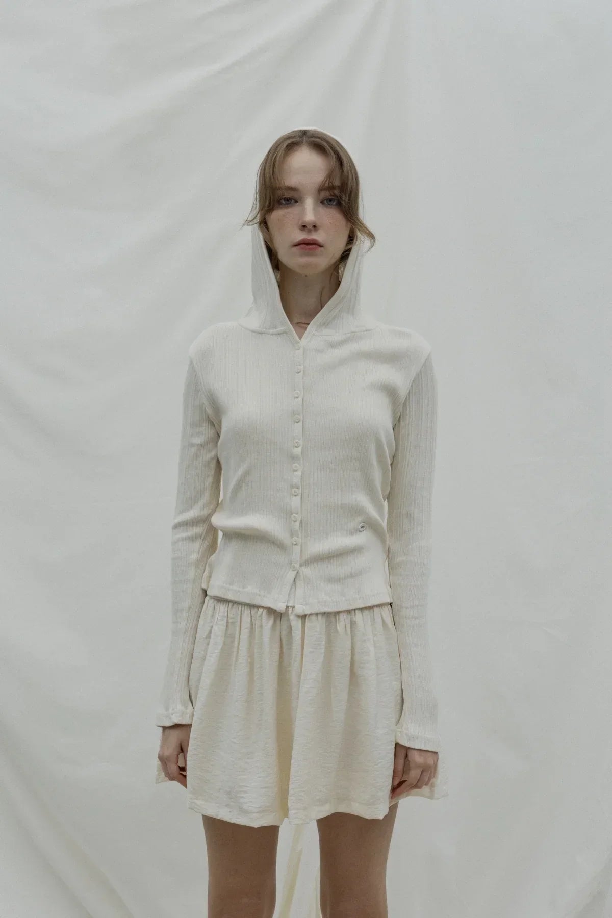Woman wearing natural cream ribbed hooded cardigan with buttons and matching skirt on plain background