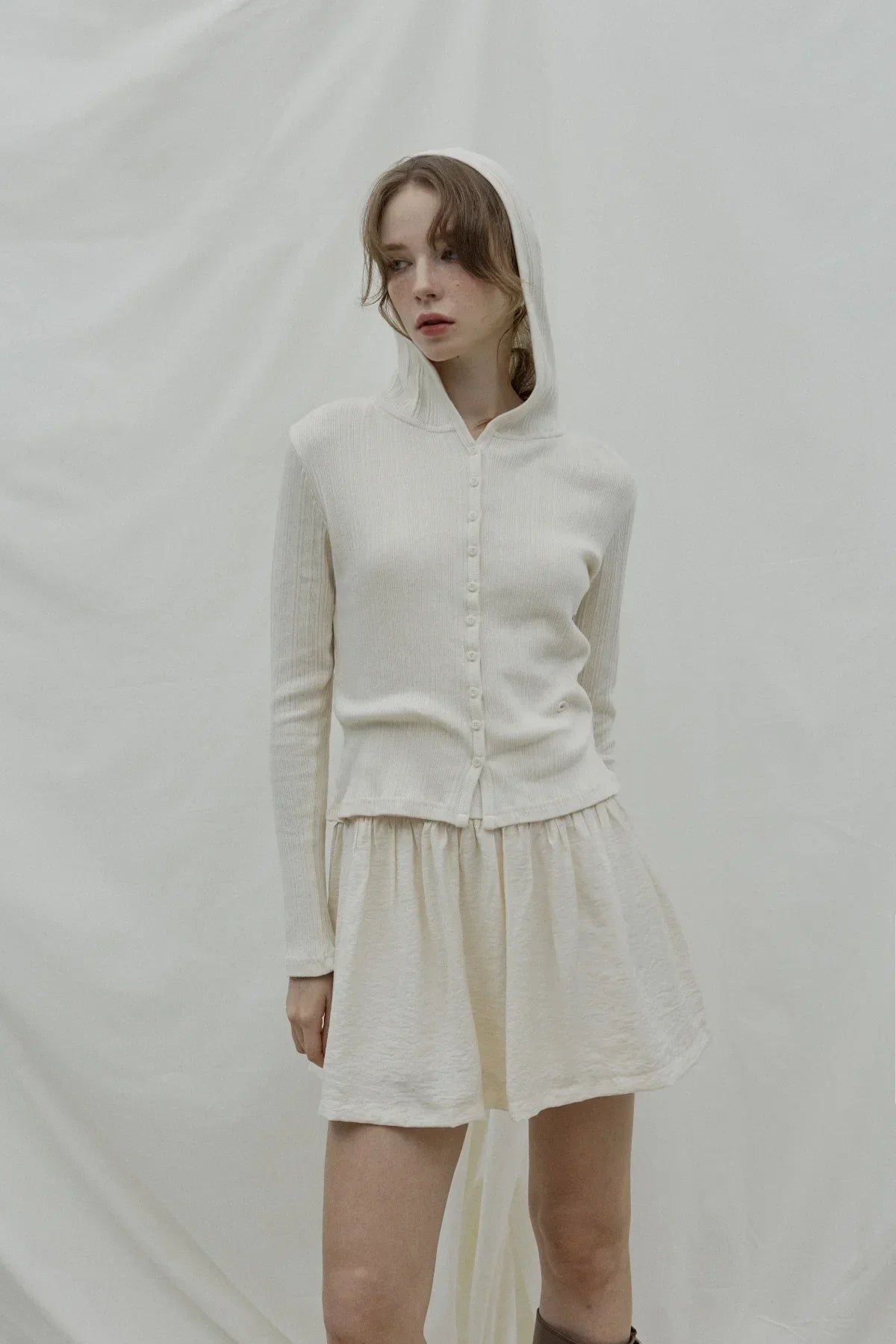 Woman wearing cream ribbed hooded cardigan with buttons and matching cream skirt against neutral backdrop