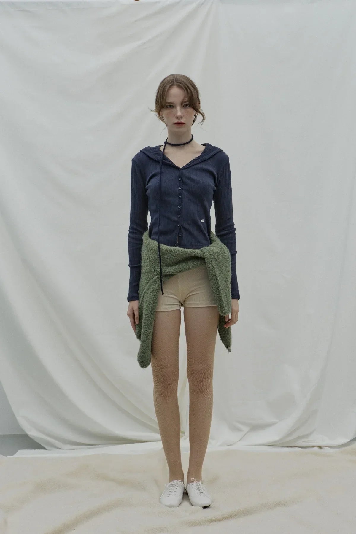 Model in navy ribbed hooded cardigan, beige shorts, tied green cardigan, white shoes on neutral backdrop