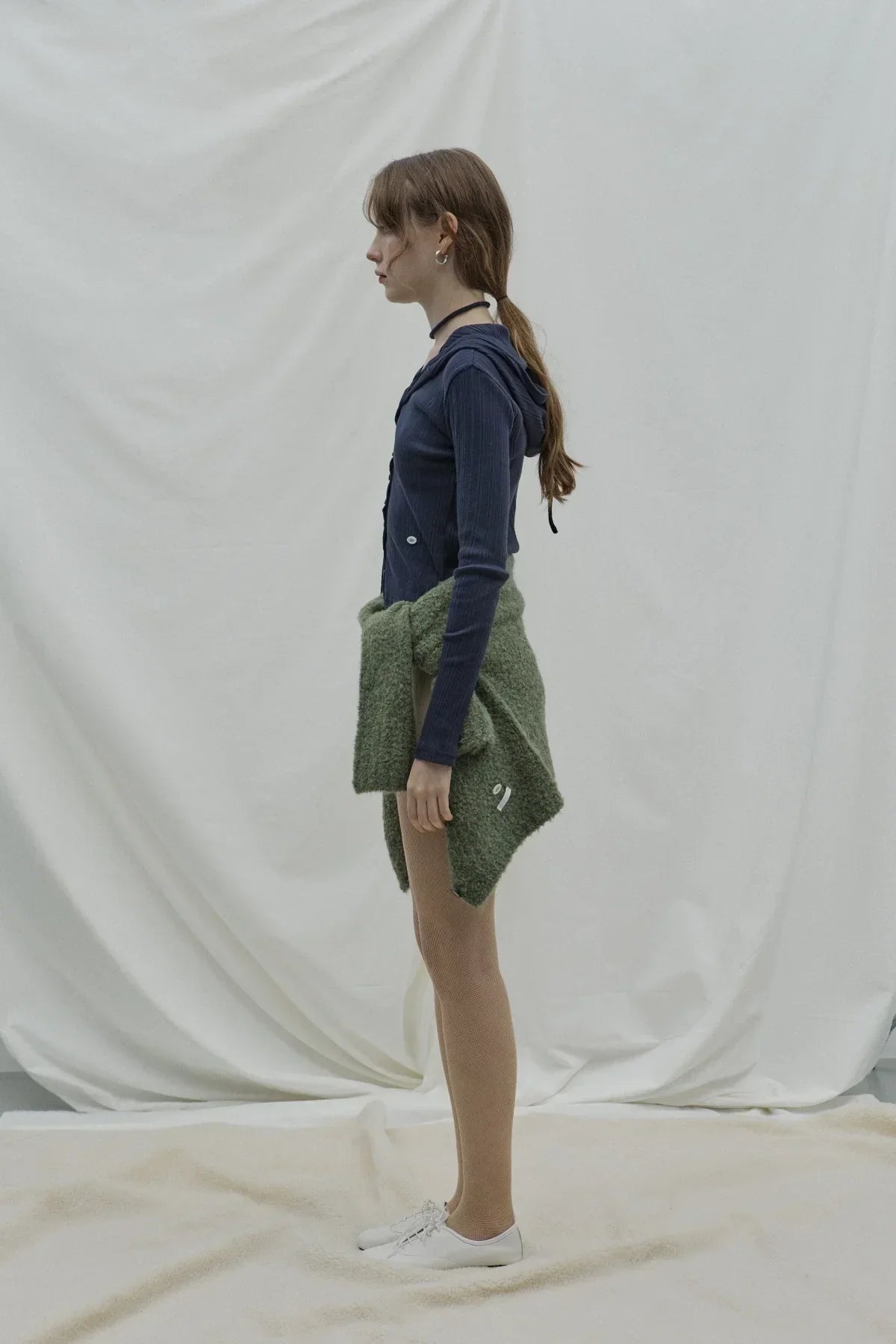 Side view of a woman wearing a navy ribbed hooded cardigan, fishnet tights, and white shoes with a green sweater tied at waist