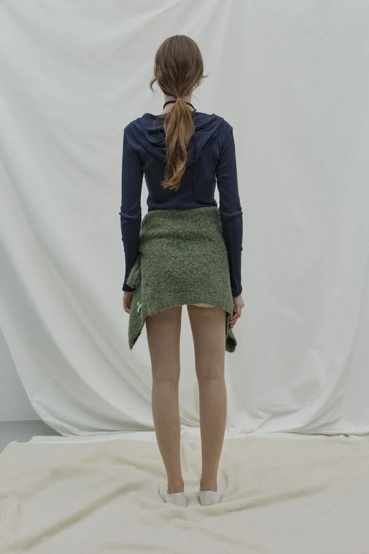 Woman wearing navy ribbed hooded cardigan with green sweater tied around waist, white shoes, standing on light fabric