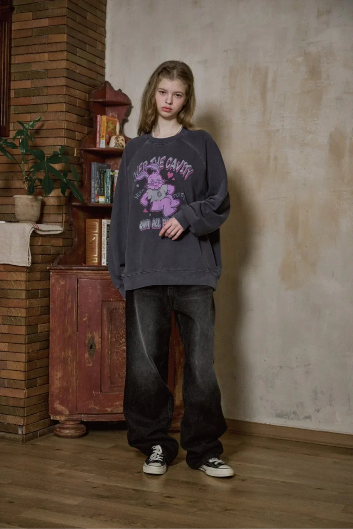 Young woman wearing dark blue oversized raglan sweater with cartoon print and baggy black jeans indoors