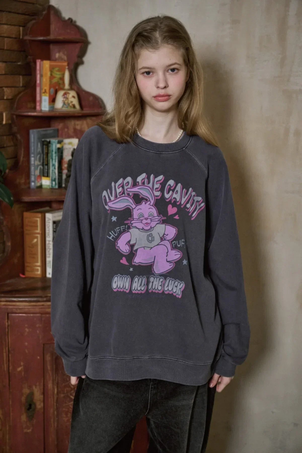 Young woman wearing dark oversized graphic sweatshirt with pink bunny and text, standing indoors by rustic bookshelf
