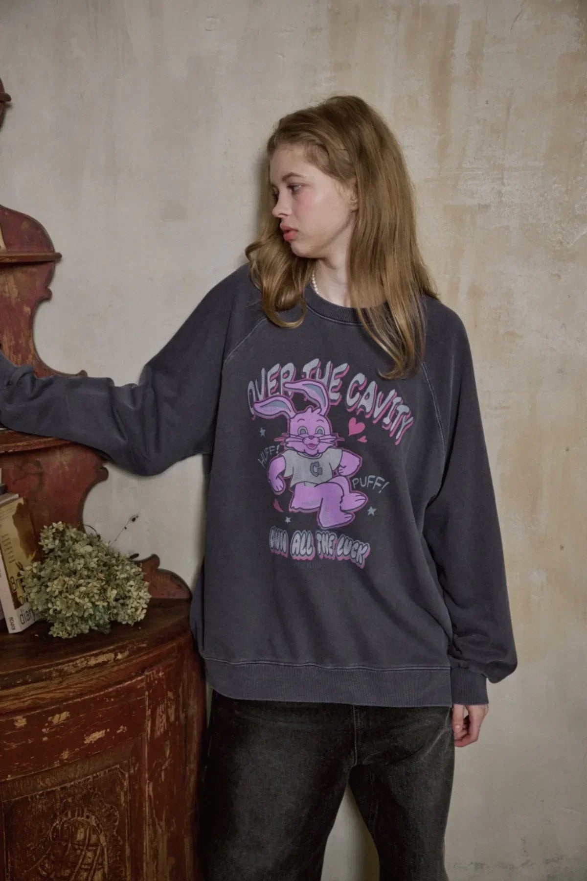 Young woman wearing dark blue oversized sweatshirt with pink cartoon bunny graphic, standing indoors by rustic wooden furniture