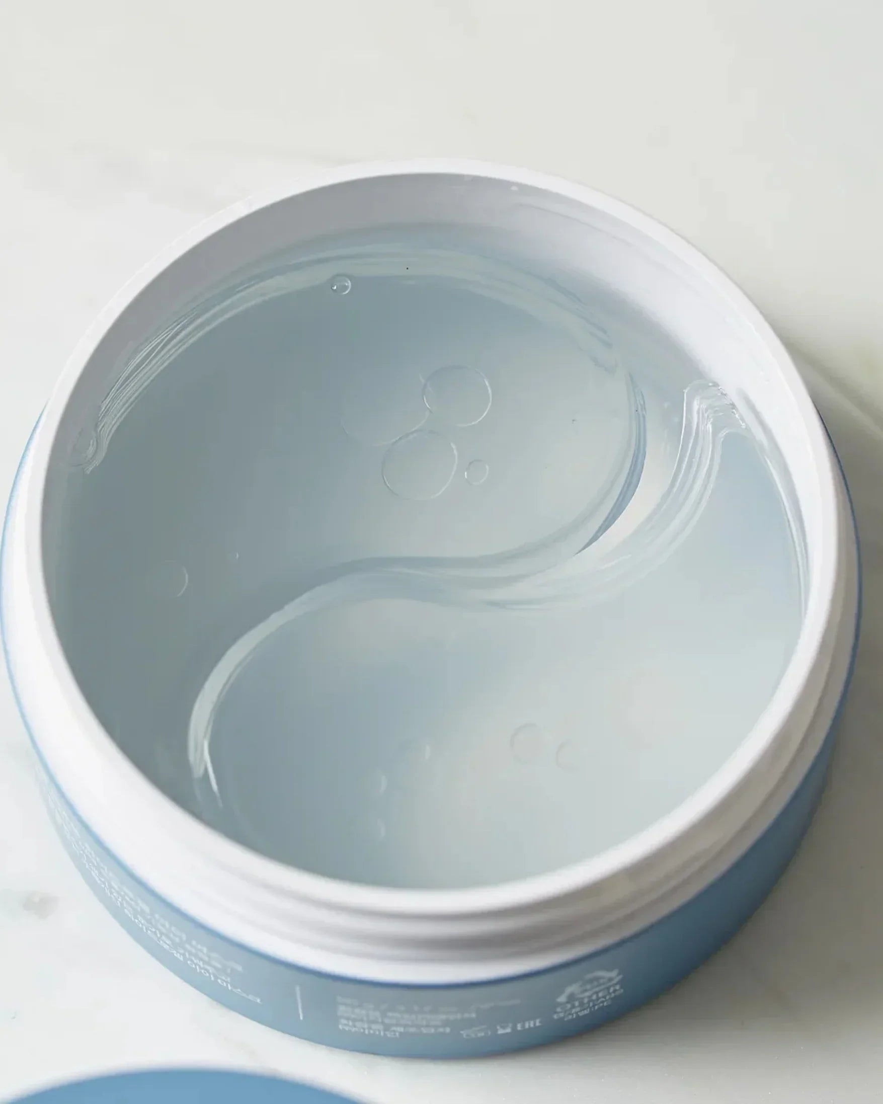 Open blue jar with clear hydrogel eye masks inside on white surface