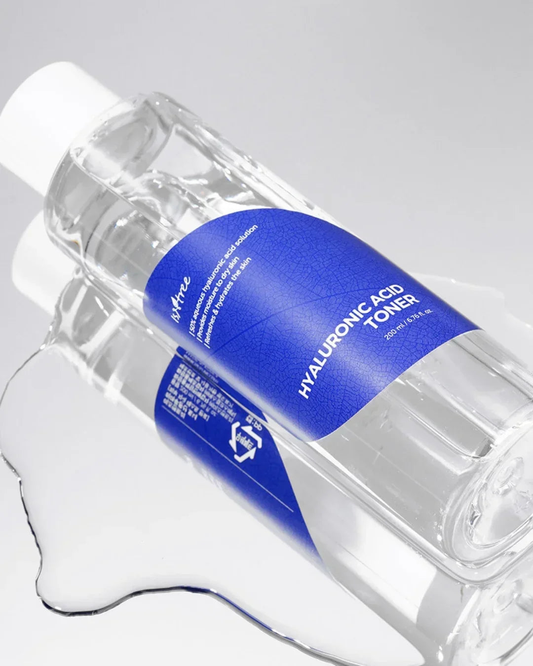 Clear plastic bottle of Hyaluronic Acid Toner with blue label lying on reflective silver surface