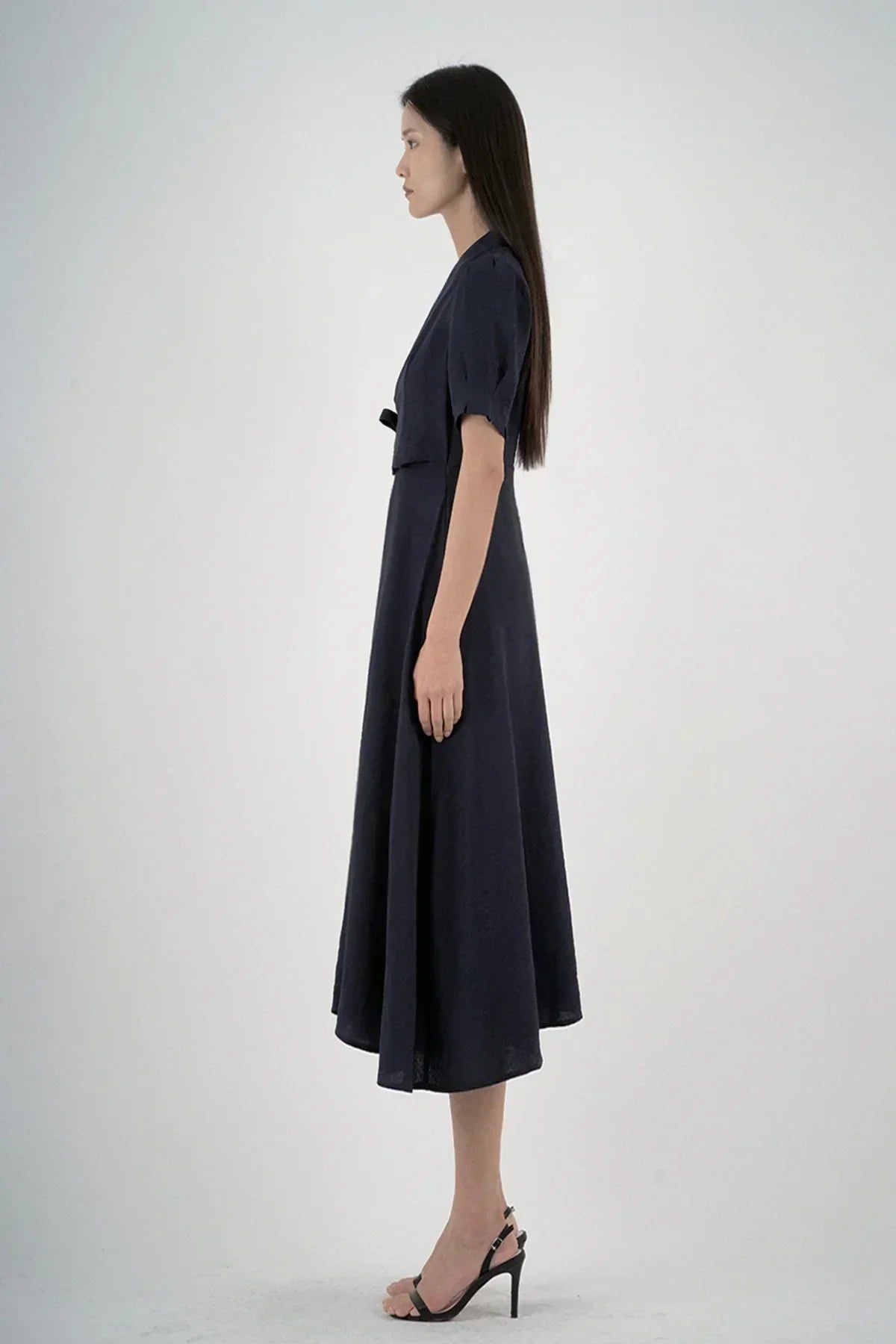 Side view of woman in navy midi dress with short sleeves and tie waist, wearing black heels on white background
