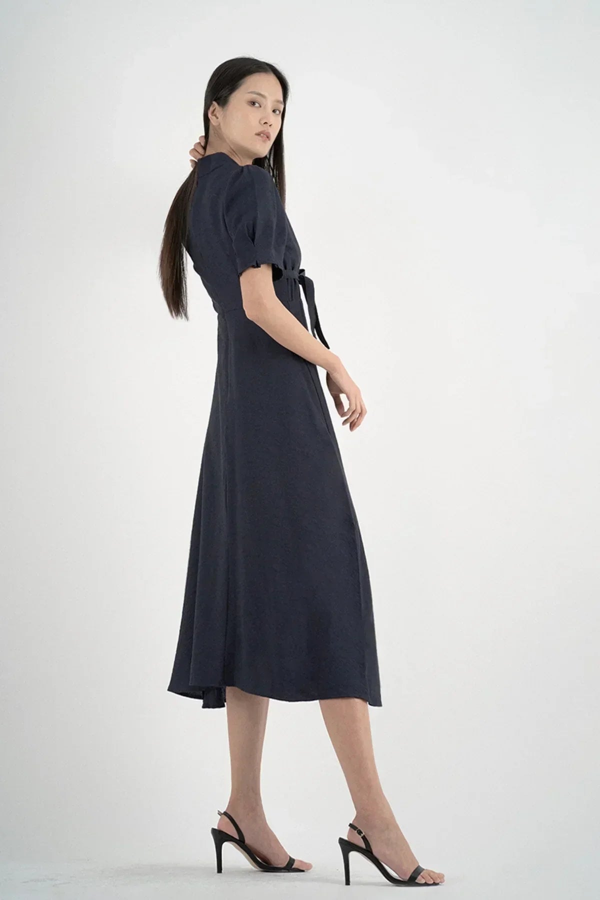 Asian woman wearing a navy blue midi dress with puff sleeves and black heels on white background