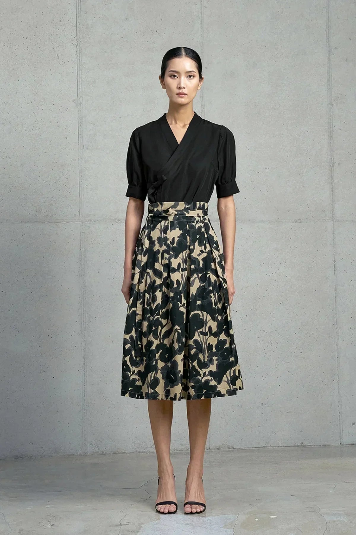 Woman wearing black wrap top and beige pleated skirt with black floral ink wash pattern standing indoors