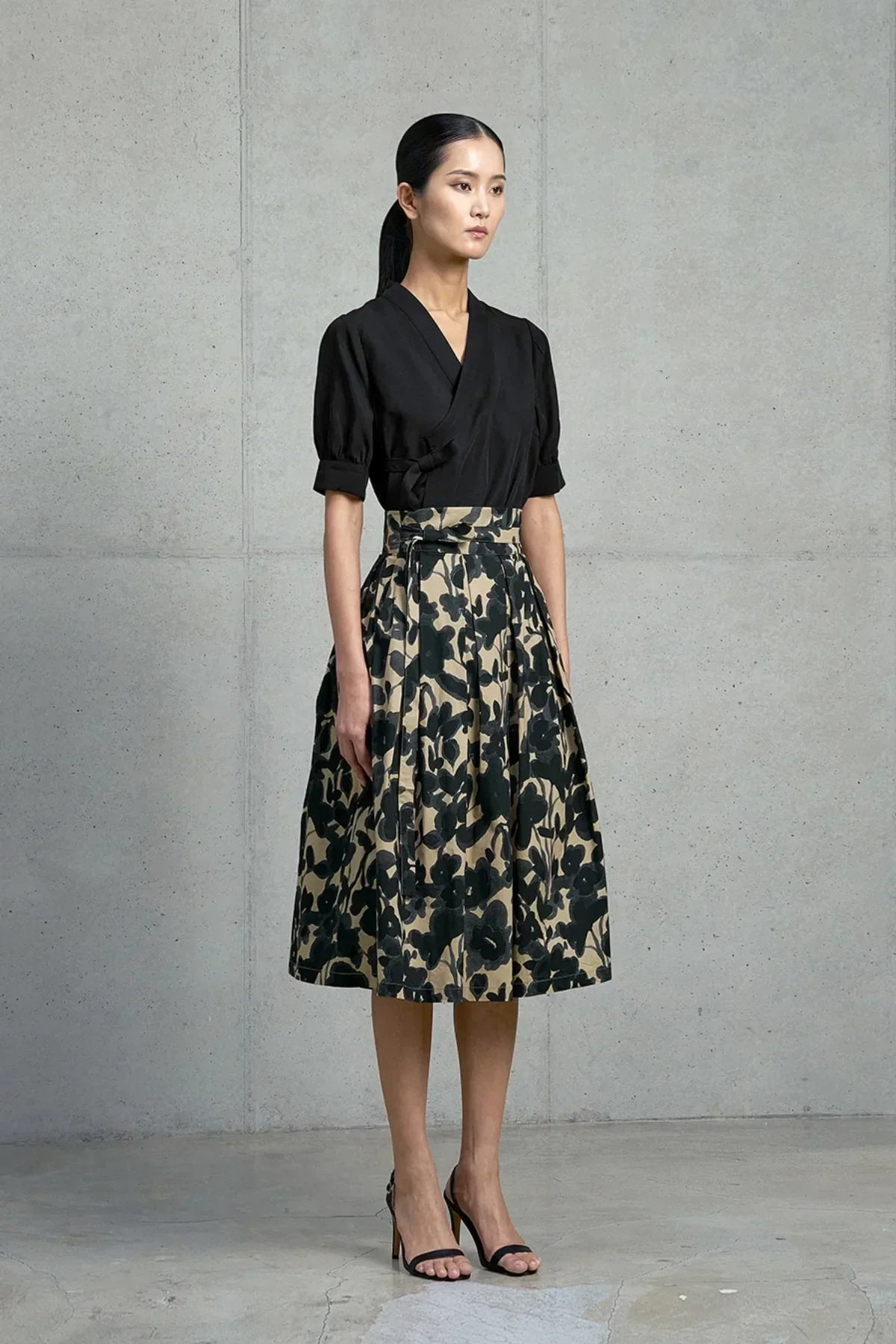 Model wearing black wrap top and beige skirt with black floral ink wash pattern, standing against gray concrete wall.