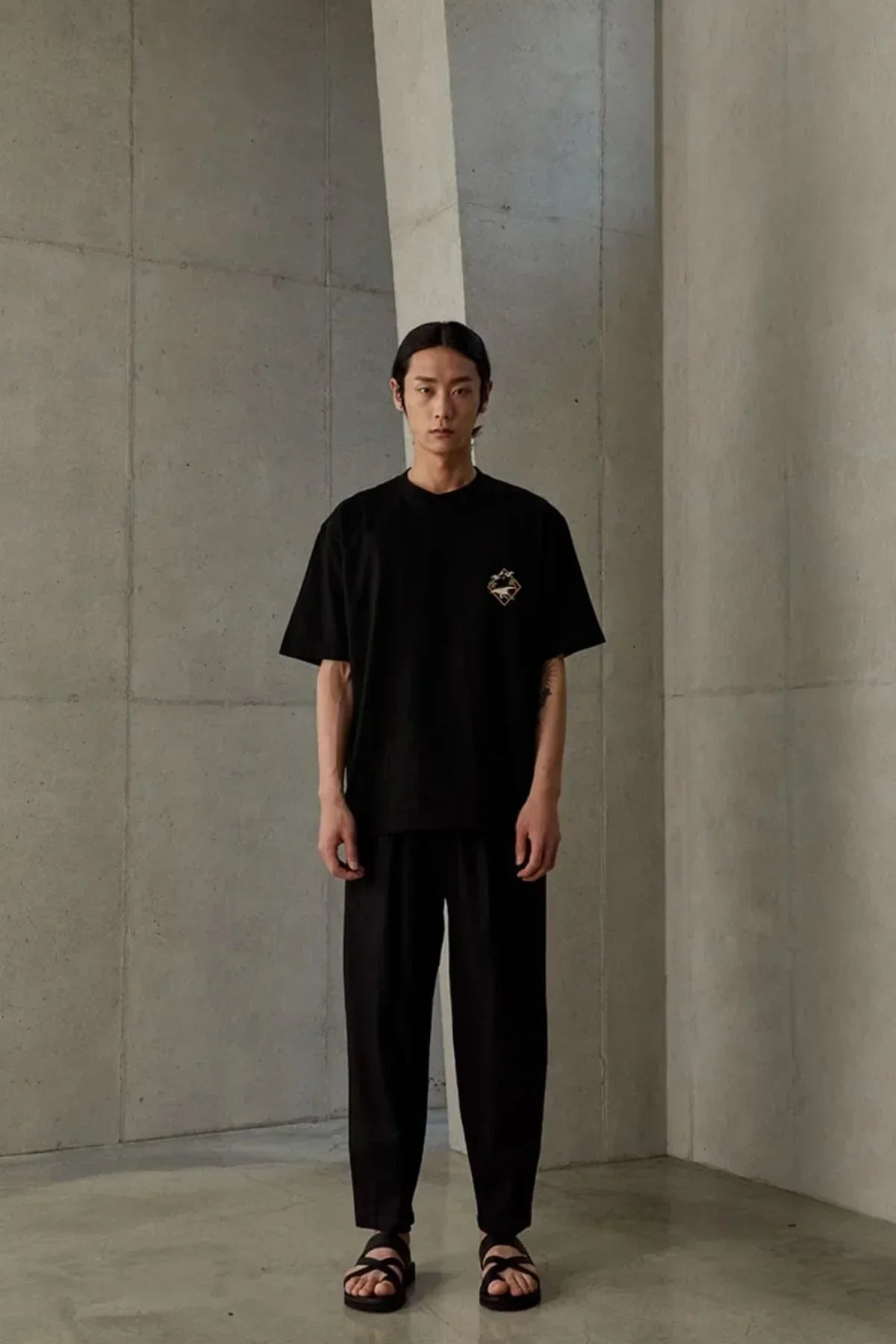 Male model wearing black oversized T-shirt with small graphic, black loose pants, and black sandals in minimalist concrete interior