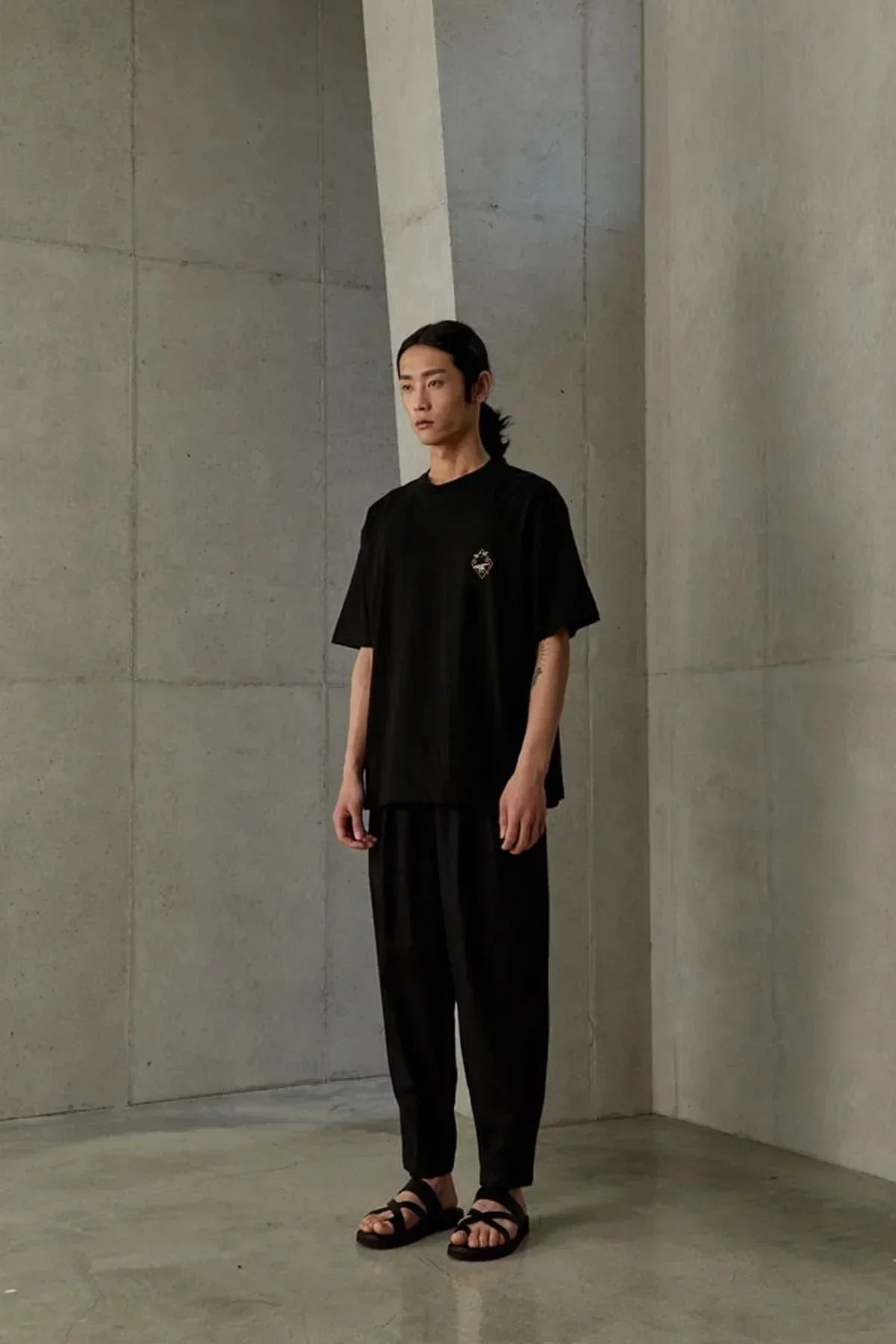 Male model wearing black jangsaengdo graphic t-shirt with loose black pants and sandals in minimalist concrete room