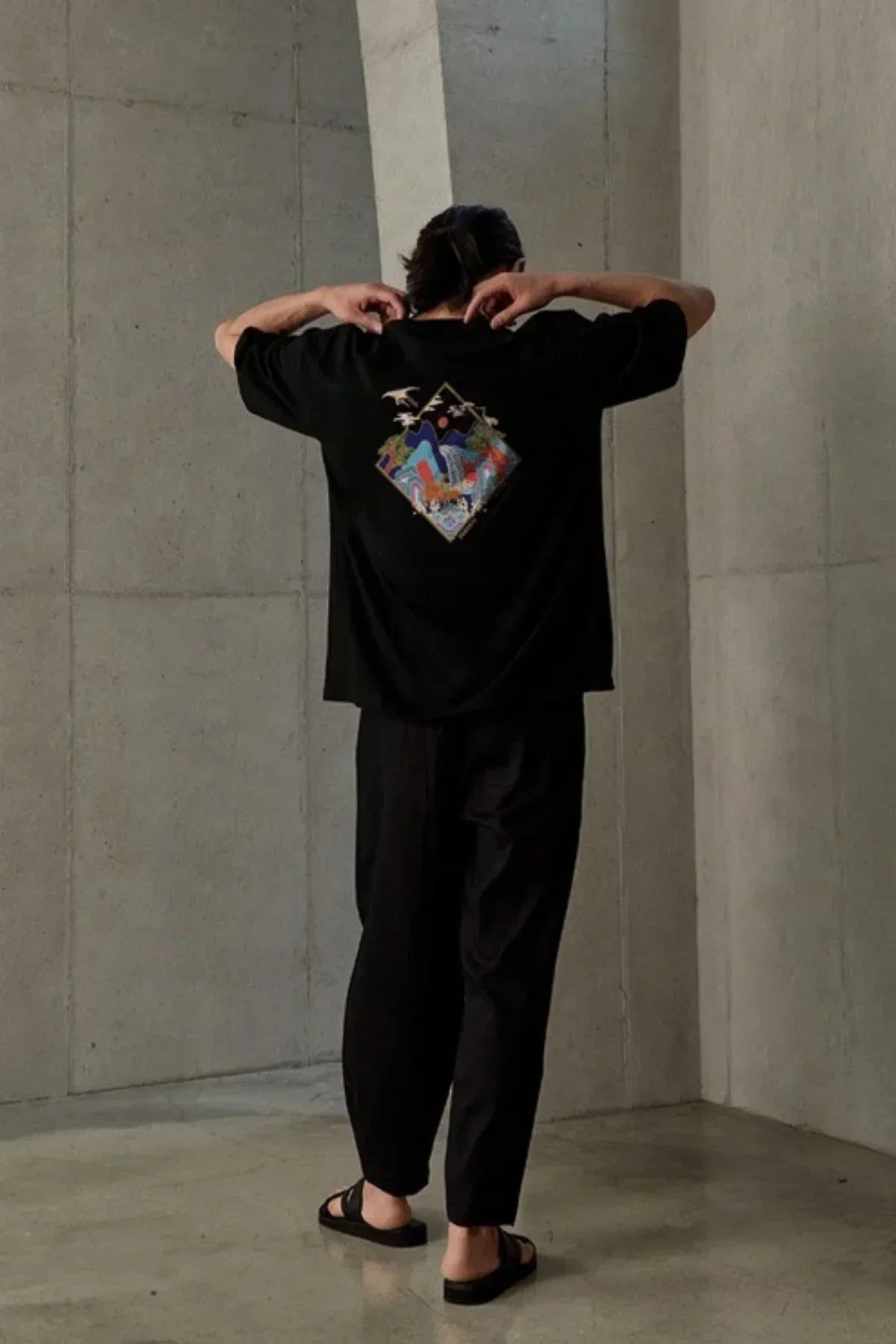 Man wearing black t-shirt with colorful abstract graphic on back, black pants, and sandals in minimalist room