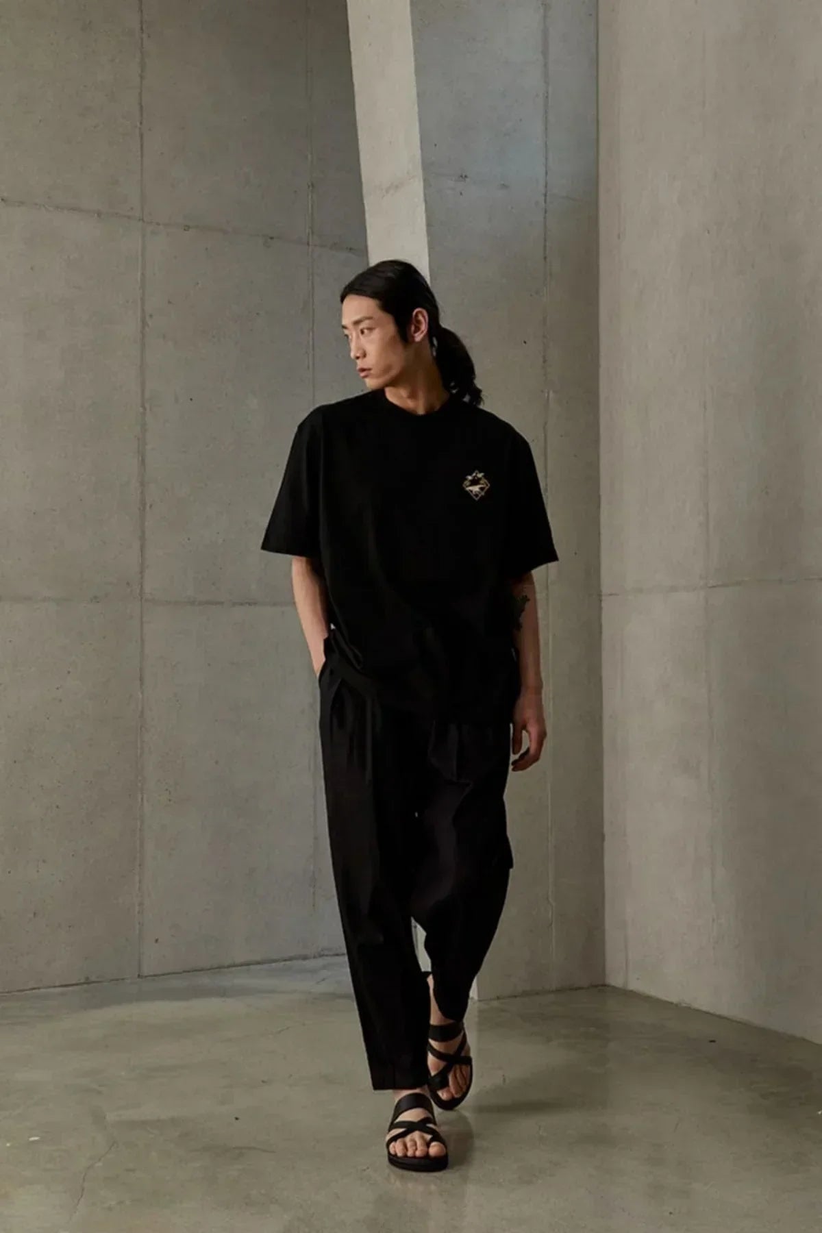 Man wearing black Jangsaengdo graphic t-shirt, black pants, and sandals in minimalist concrete room