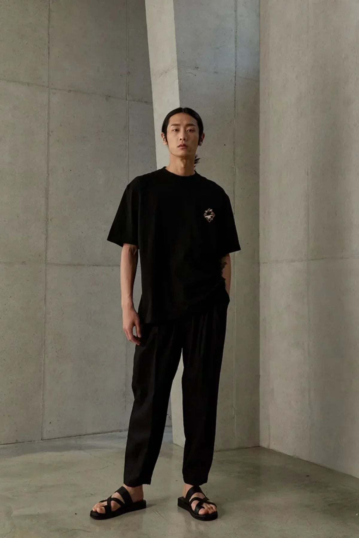 Man wearing black jangsaengdo graphic t-shirt, black pants, and sandals standing in minimalist concrete studio