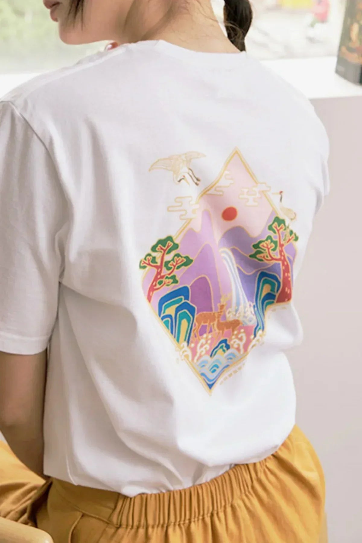 White t-shirt with colorful mountain, trees, deer, and crane graphic worn by person in mustard pants