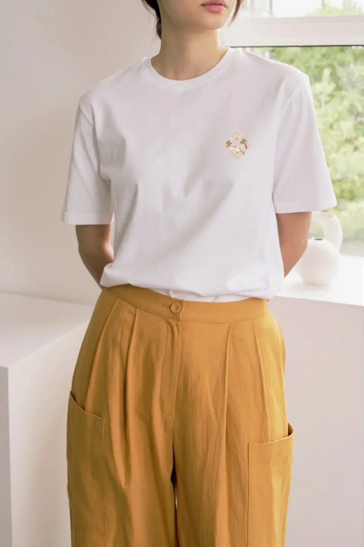 Woman wearing white t-shirt with small pink and gold graphic paired with mustard yellow pants indoors