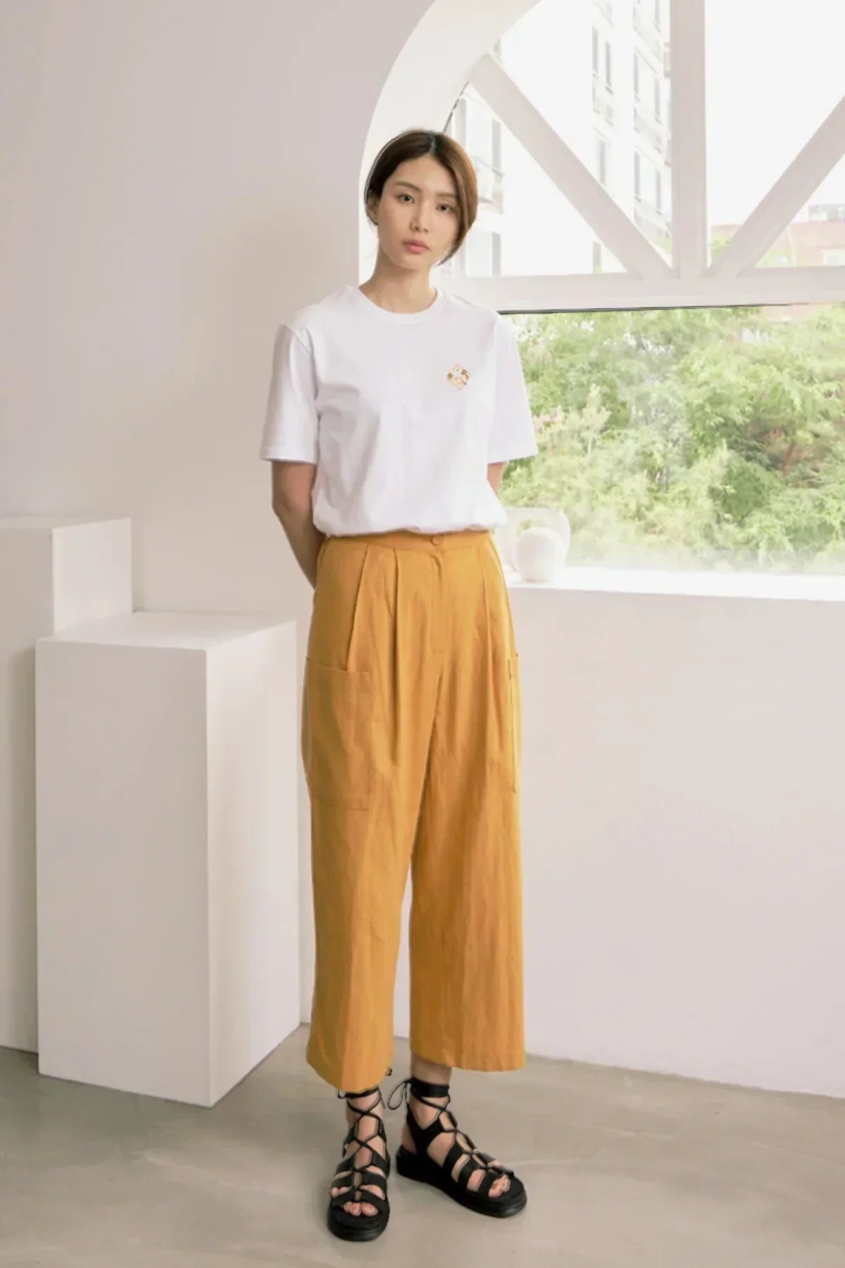 Woman in white graphic t-shirt, mustard wide-leg pants, and black gladiator sandals in modern indoor setting