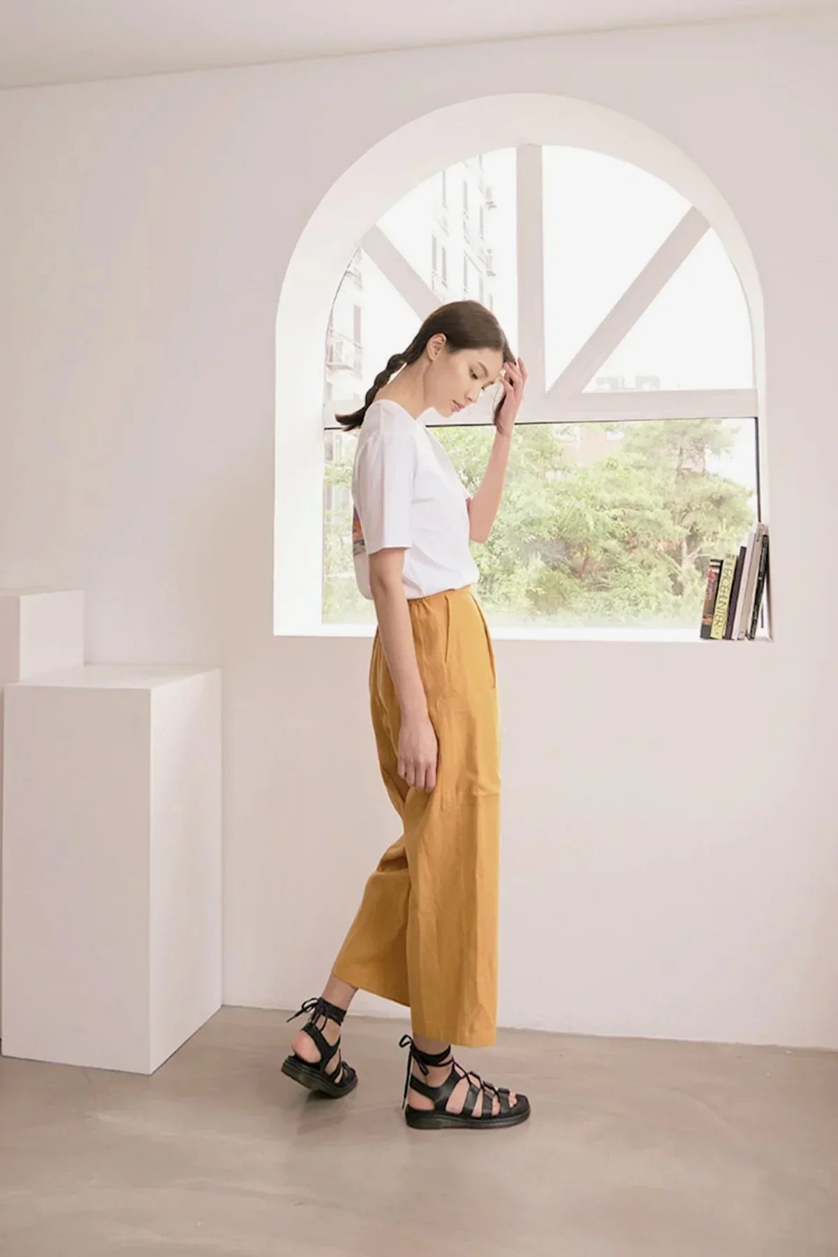 Woman in white t-shirt and mustard pants wearing black sandals standing in minimalist room with arched window