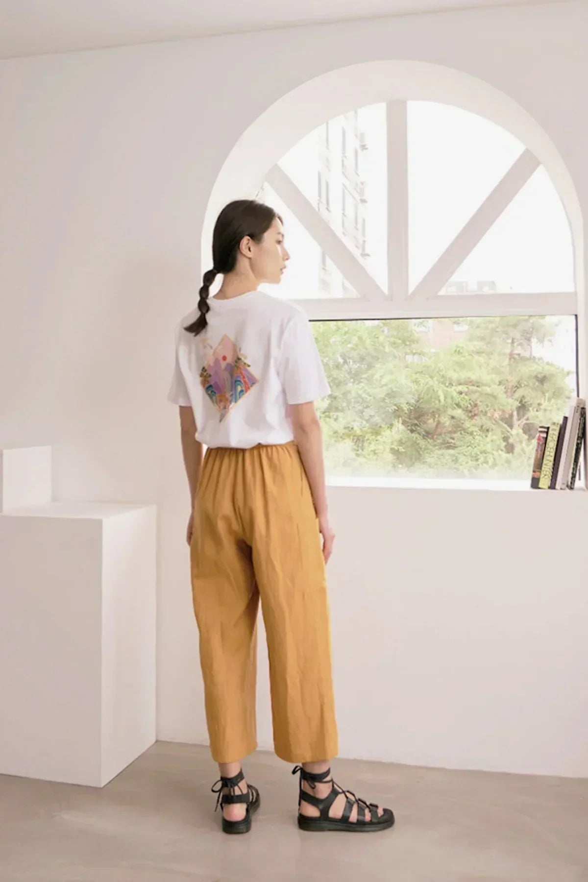 Woman wearing white graphic t-shirt and mustard wide-leg pants standing by an arched window with books