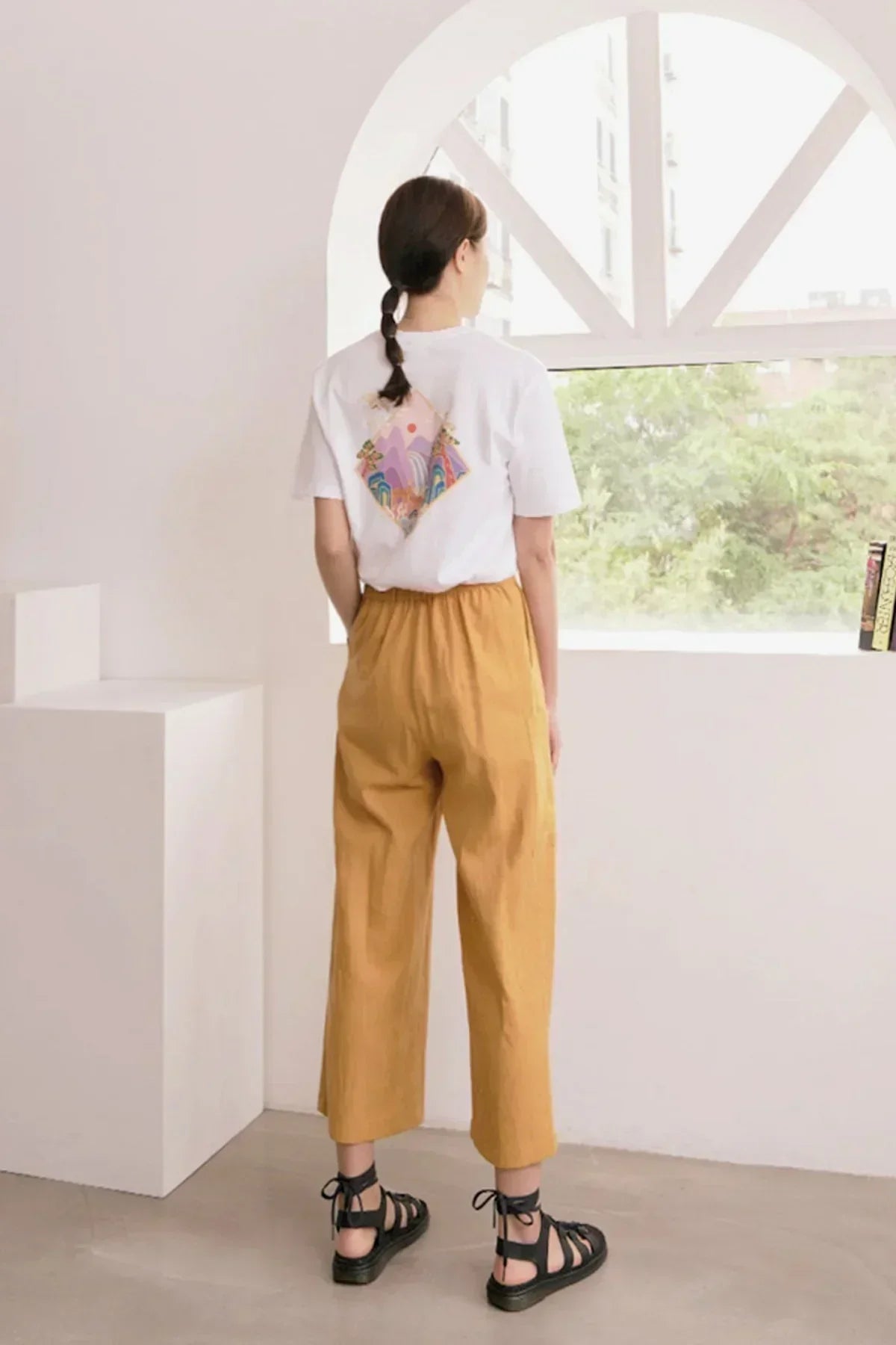 Woman in white graphic t-shirt and mustard wide-leg pants standing by arched window in minimalist room