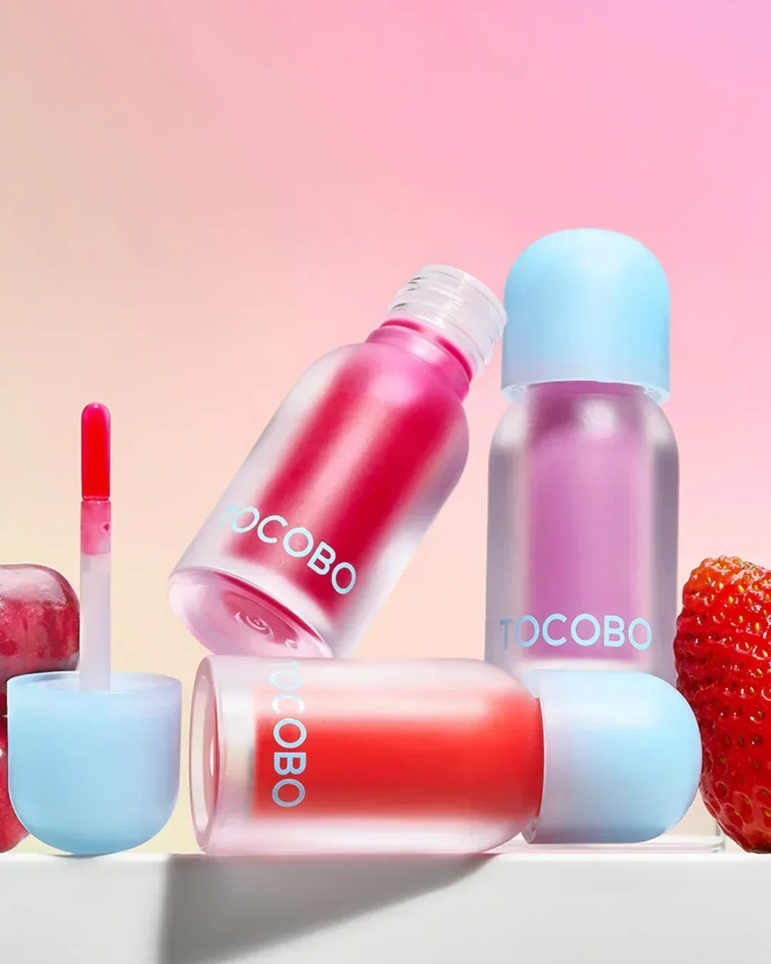 TOCOBO juicy berry plumping lip oil in frosted bottles with blue caps and strawberry backdrop